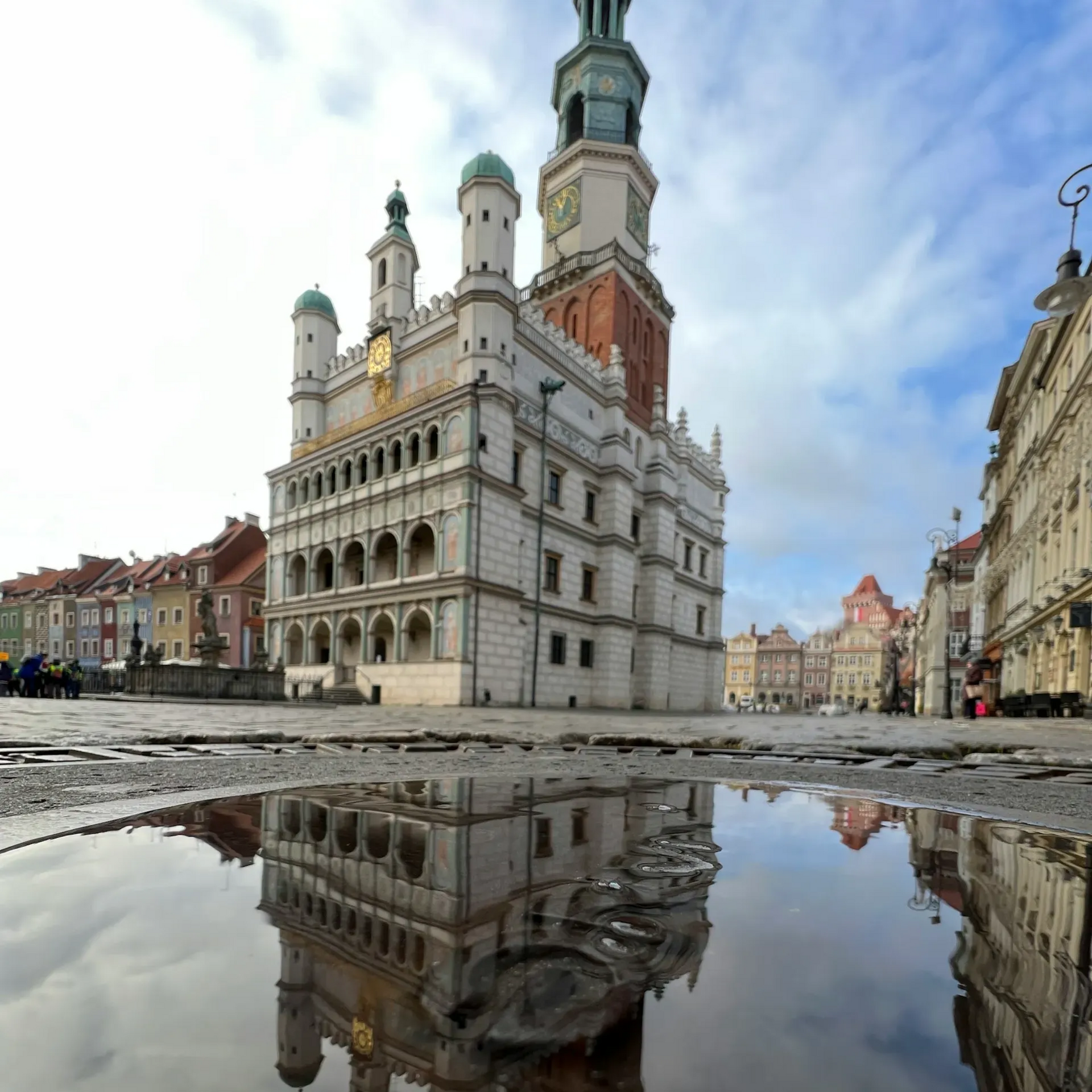 Budynek Ratusza odbija się w kałuży na brukowanym placu w Poznaniu. Nad głową zachmurzone niebo.