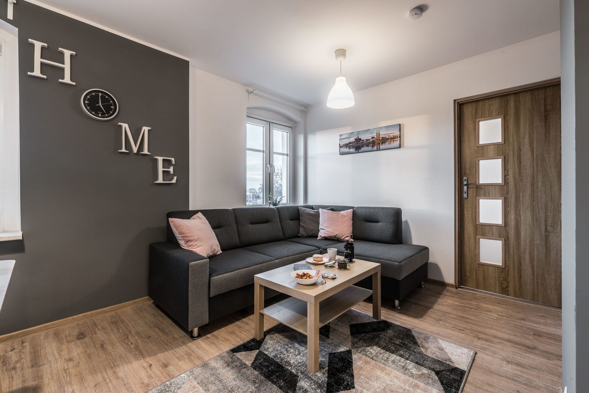 Salon z szarą narożną sofą, dekoracją ścienną z napisem „HOME” i małym stolikiem kawowym.
