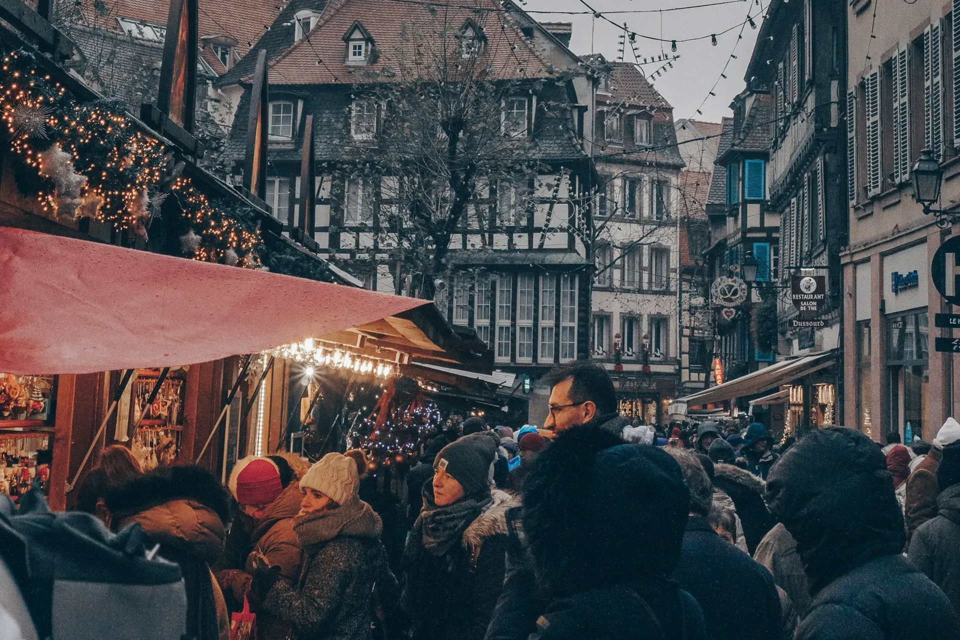 Crowd at a European Christmas market with stalls, lights, and timber-framed buildings.