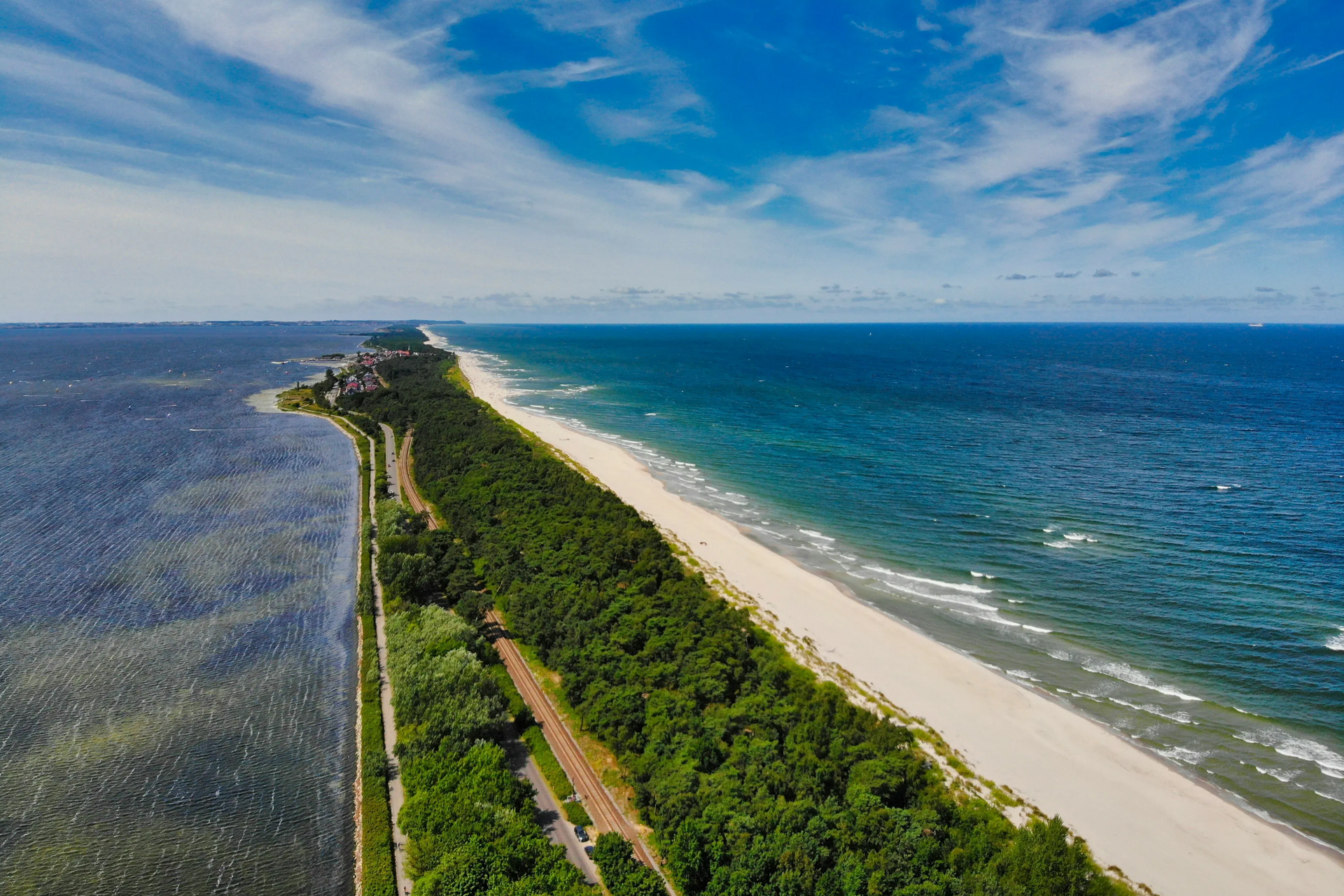Widok z lotu ptaka na wąski pas lądu porośnięty lasem i piaszczystą plażą oddzielającą morze od laguny, pod błękitnym niebem.