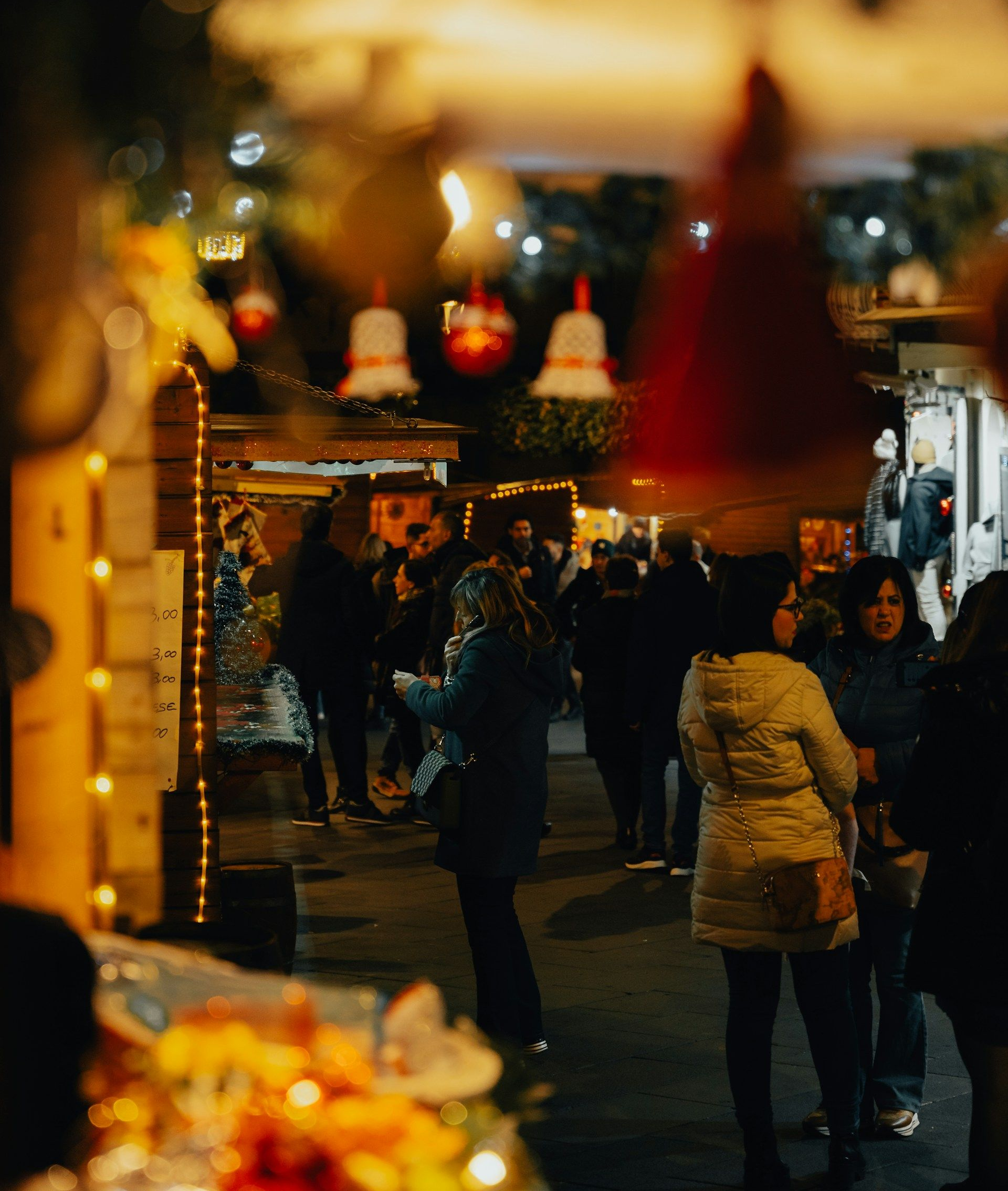 Weihnachtsmarkt bei Nacht, Menschenmenge, Lichterketten, Glockenschmuck und Stände mit beleuchteten Dekorationen.