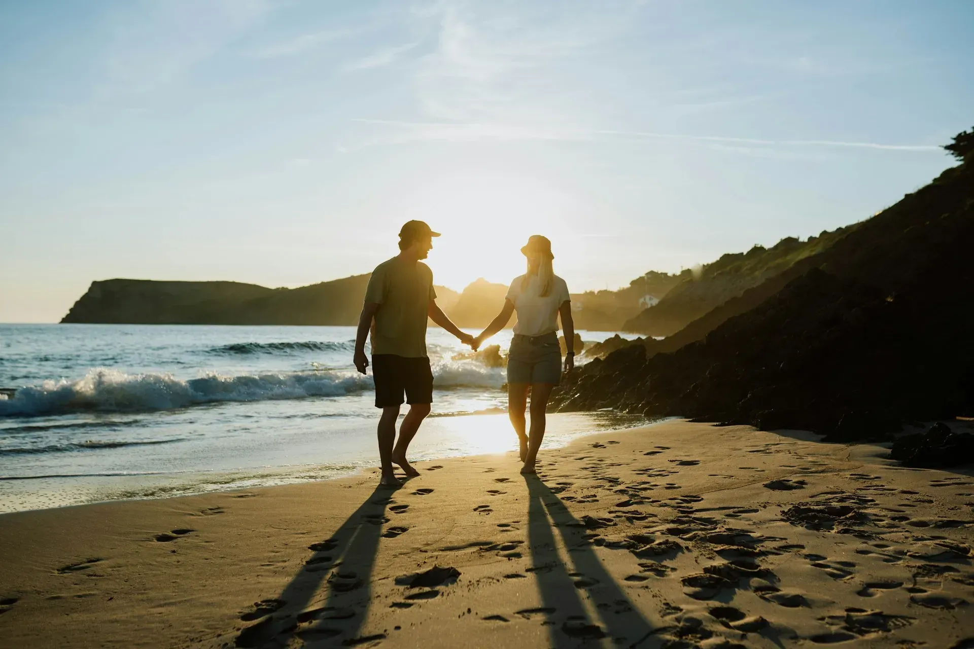 Couple holding hands, walking on a beach at sunset, with ocean and cliffs in the background.