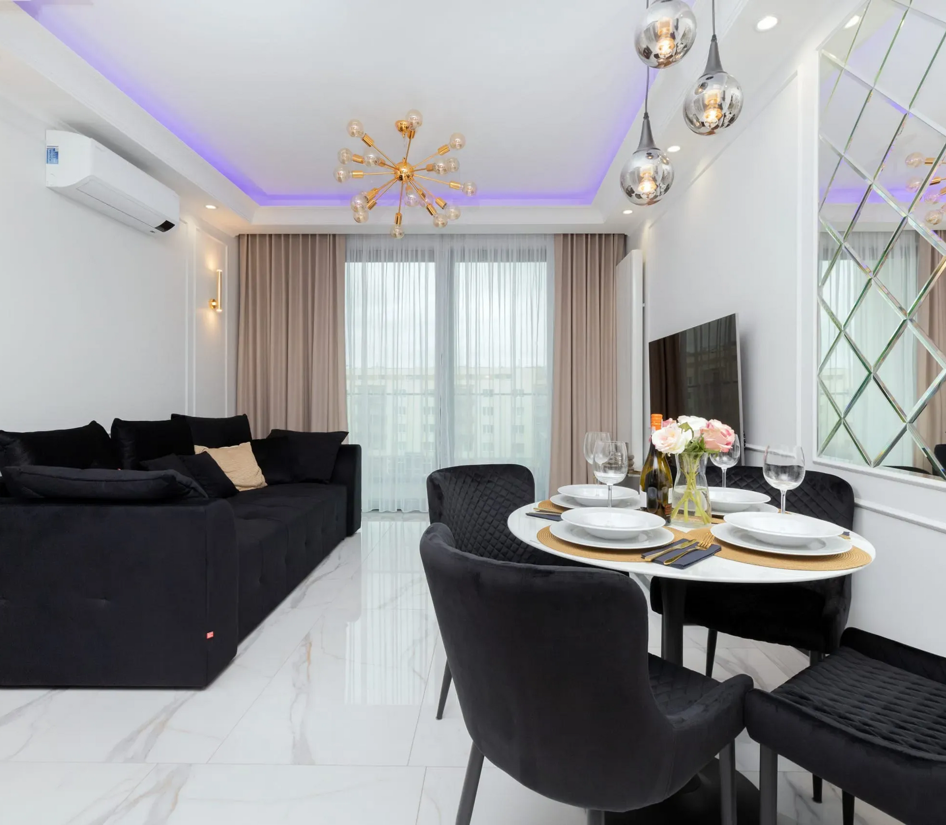 Salon z czarną sofą, stołem jadalnym i dużym oknem. Białe ściany, złoty żyrandol i kwiatowy element dekoracyjny.