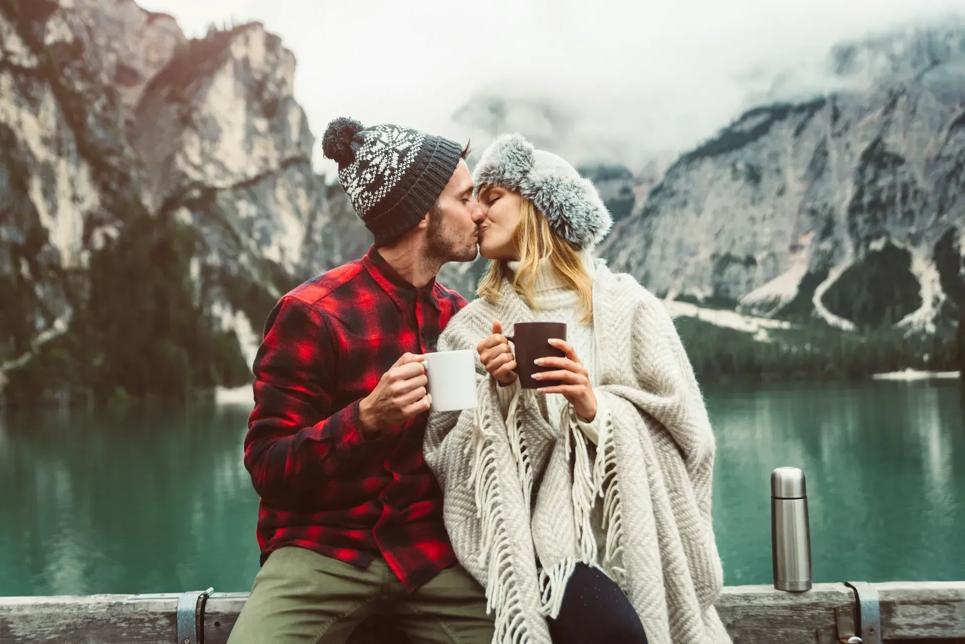 Couple kissing outdoors near lake and mountains; man holds mug, woman holds phone, wrapped in blanket.