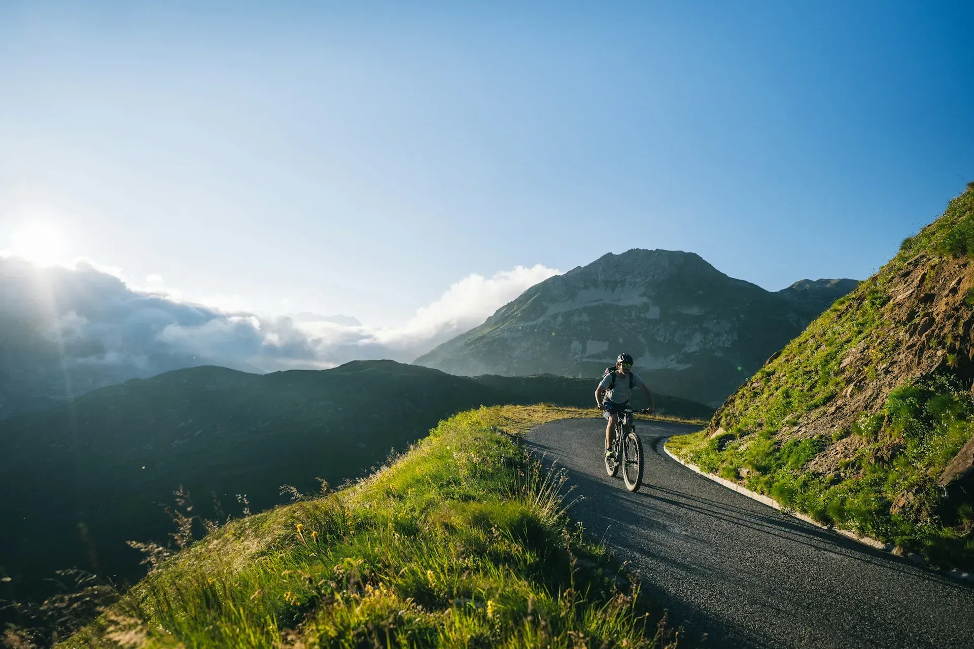 Cyclist rides uphill on a mountain road with a view of mountains and the sky.