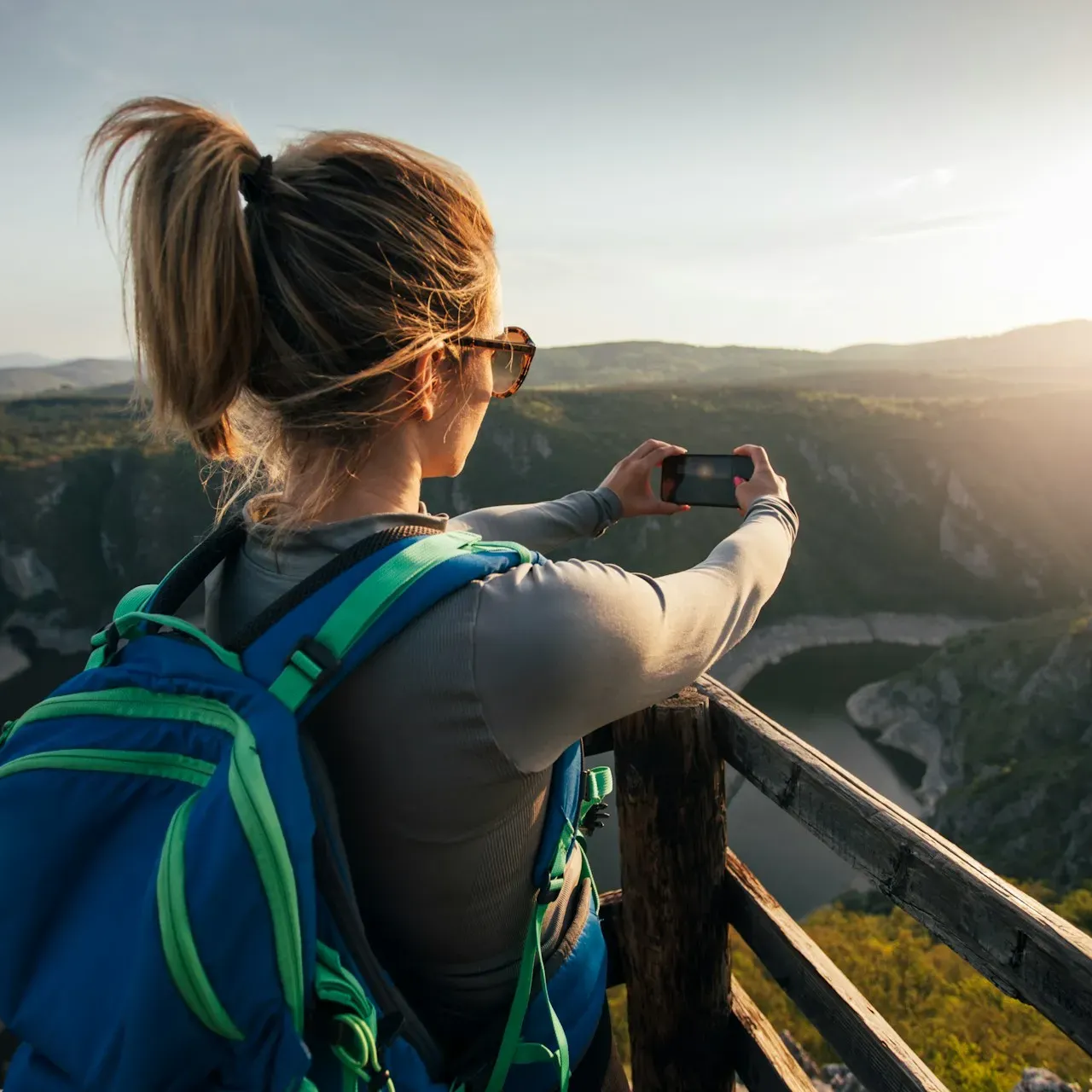 Kobieta z kucykiem, okularami przeciwsłonecznymi i plecakiem fotografuje telefonem malowniczy widok z drewnianego punktu widokowego o zachodzie słońca.