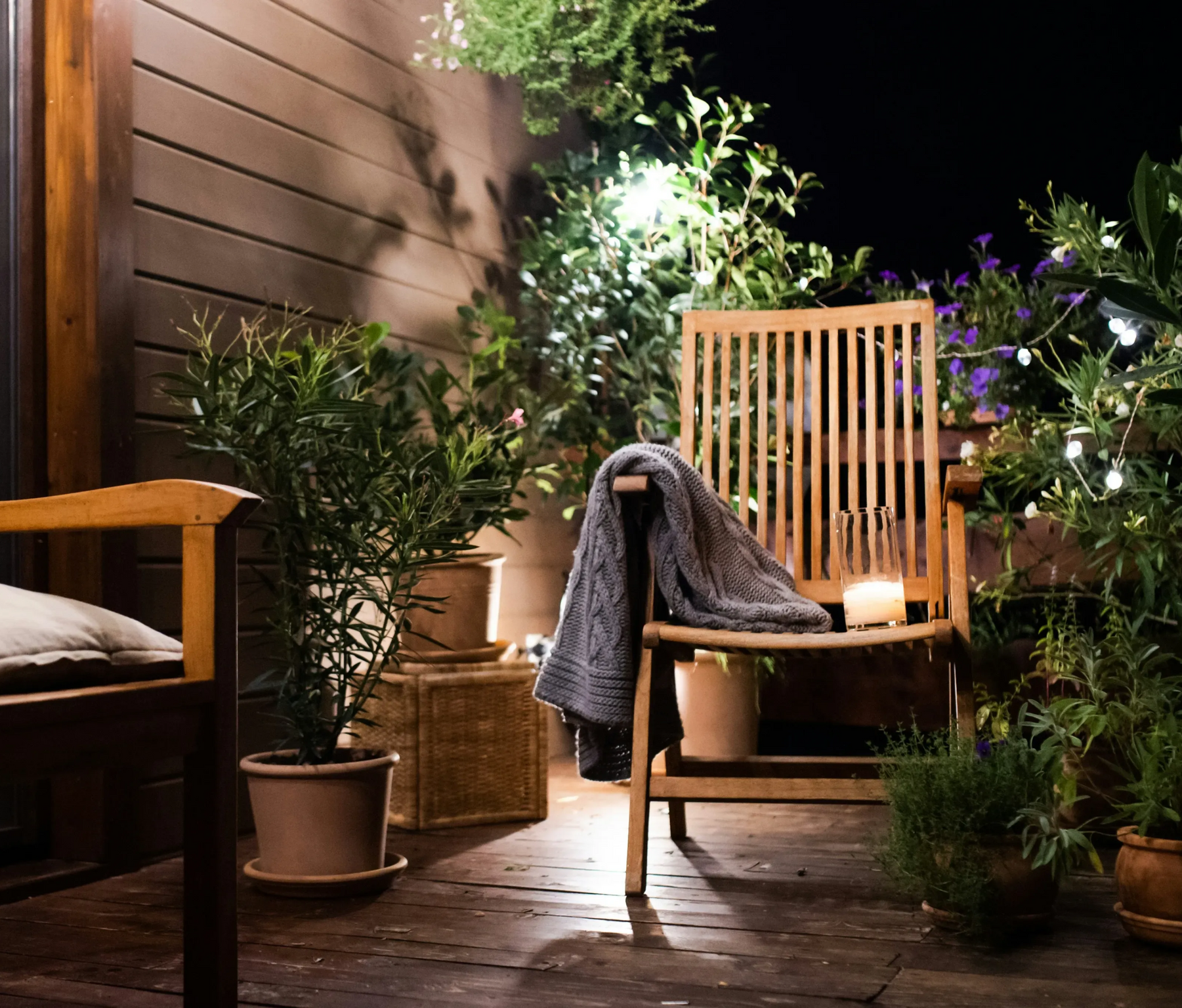 Drewniane krzesło na tarasie nocą, koc rozłożony na siedzisku, otoczenie doniczkowych roślin i delikatne oświetlenie.