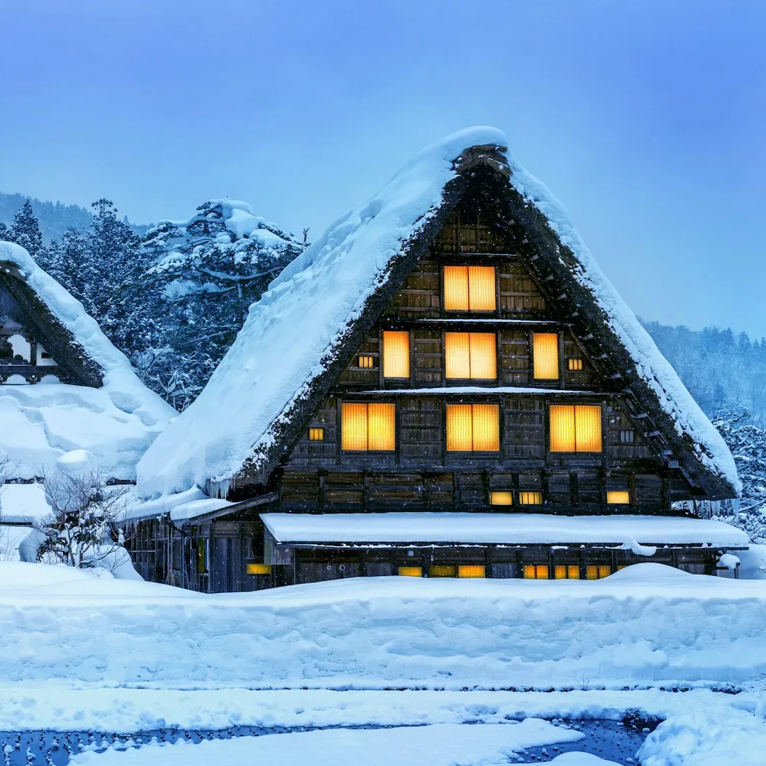 Pokryty śniegiem dom w stylu gassho w zimowym krajobrazie, oświetlone okna emanują ciepłym blaskiem.