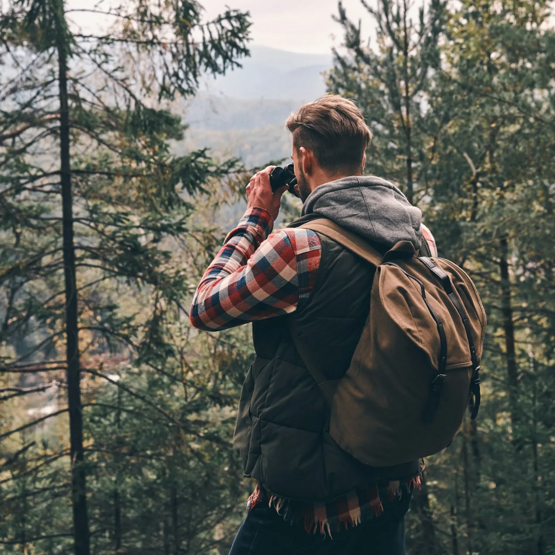 Mężczyzna z plecakiem w lesie, używa lornetki, aby obserwować górski krajobraz.
