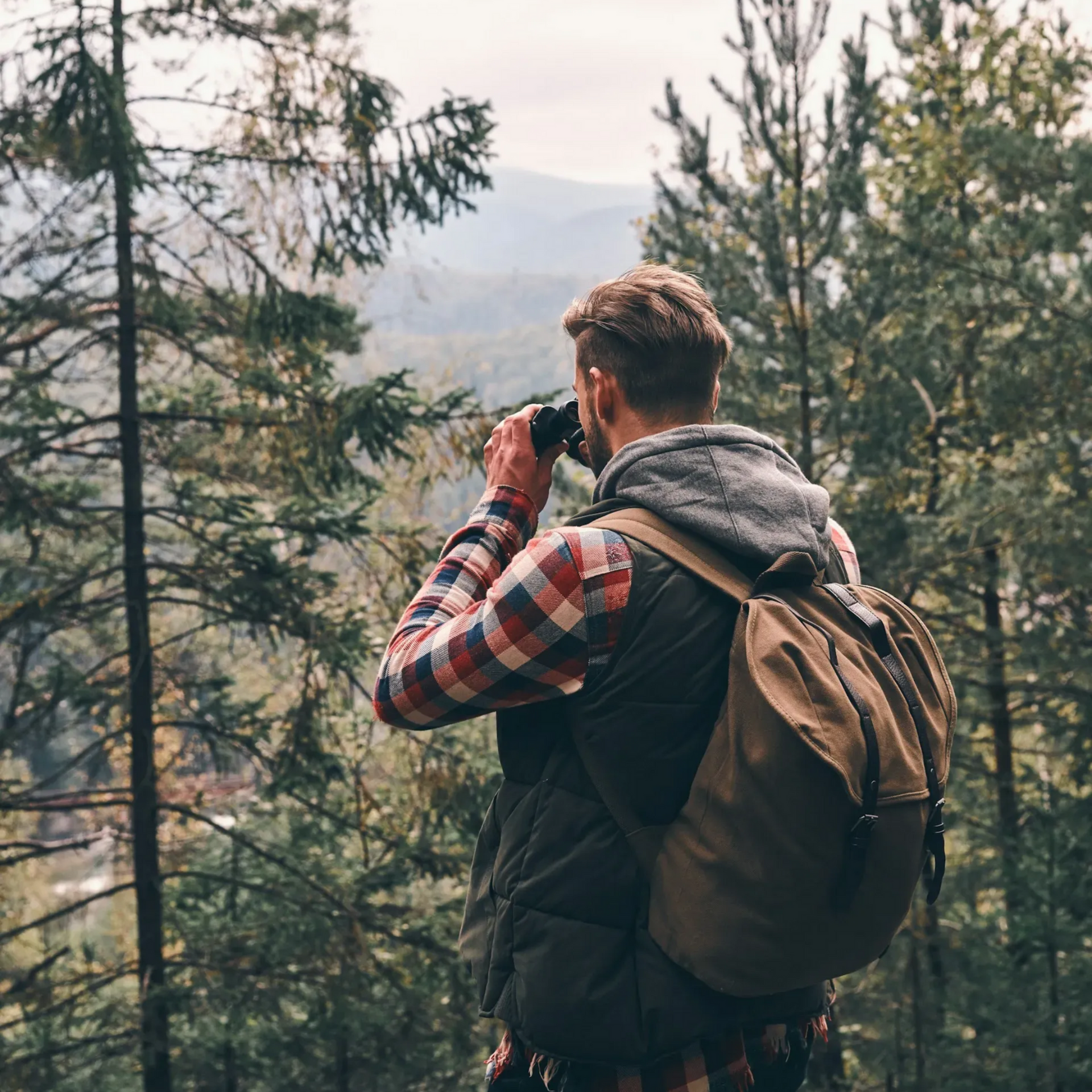 Mann mit Rucksack macht ein Foto in einem Wald, trägt eine Weste und ein kariertes Hemd.