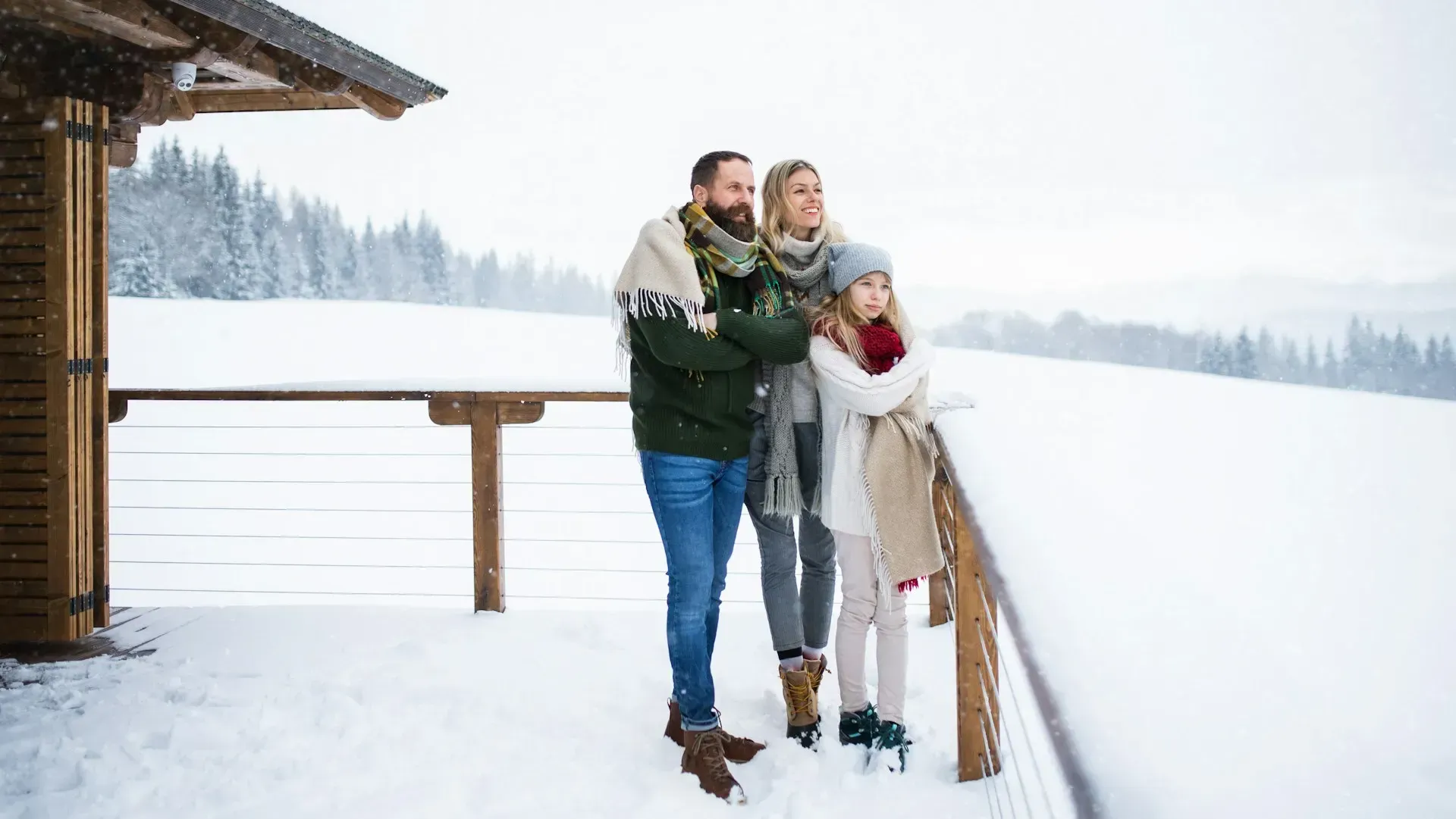 Family bundled in warm clothes on a snowy balcony, overlooking a snow-covered landscape.