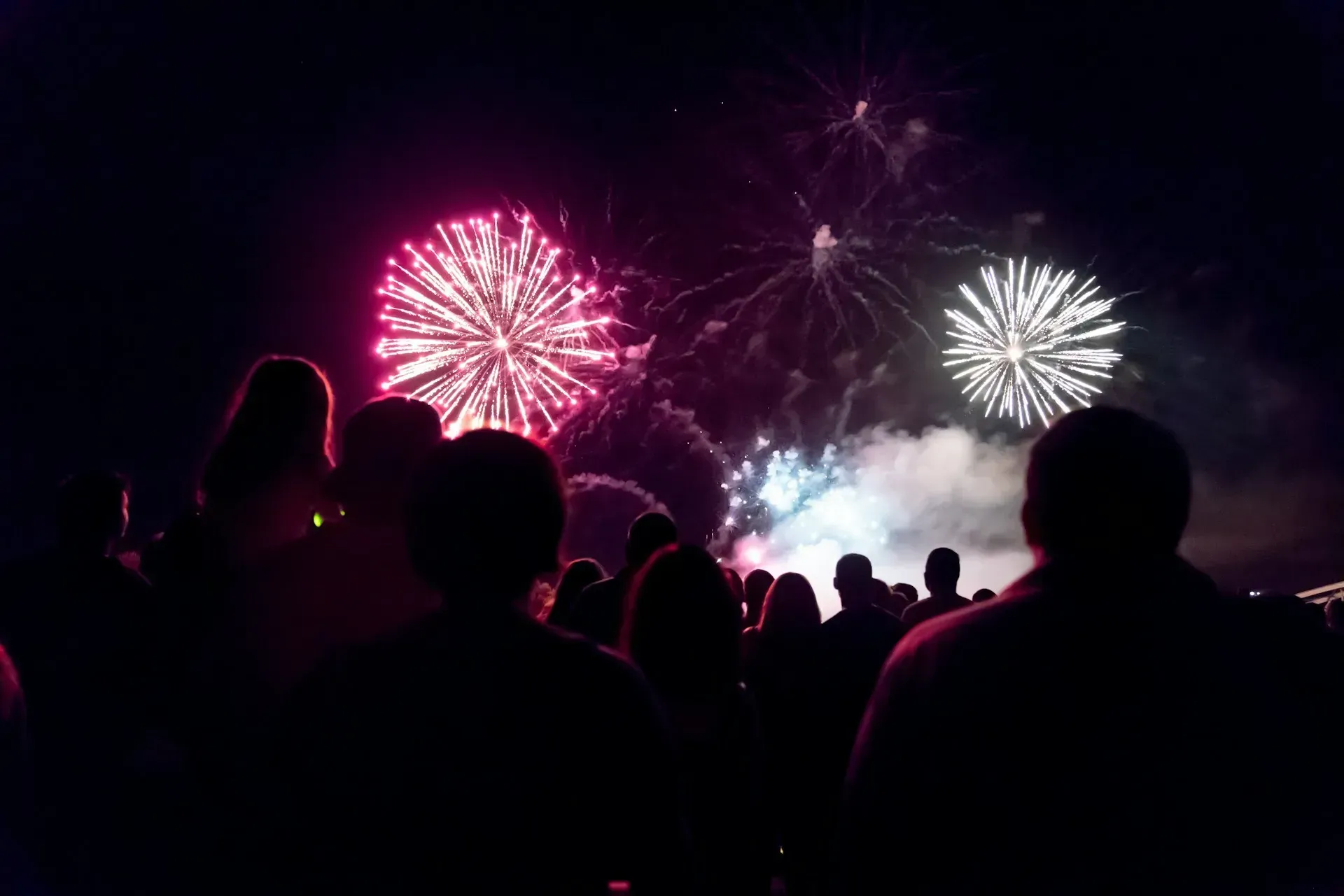 People watch fireworks explode in the night sky, illuminated by pink and white bursts.