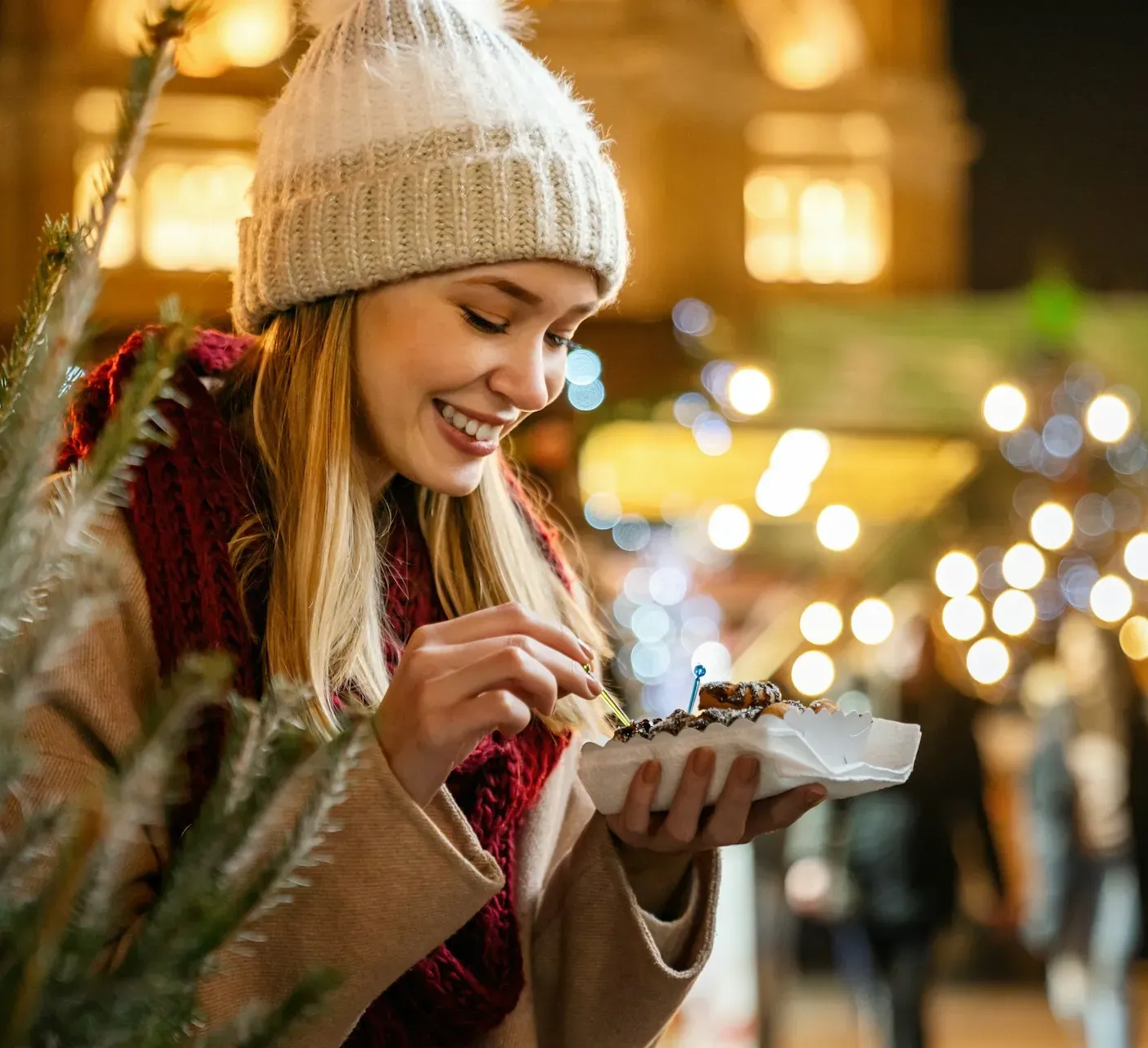 Kobieta na jarmarku je ciastka i uśmiecha się. Ma na sobie czapkę, szalik i płaszcz. W tle widać światła i budynki.