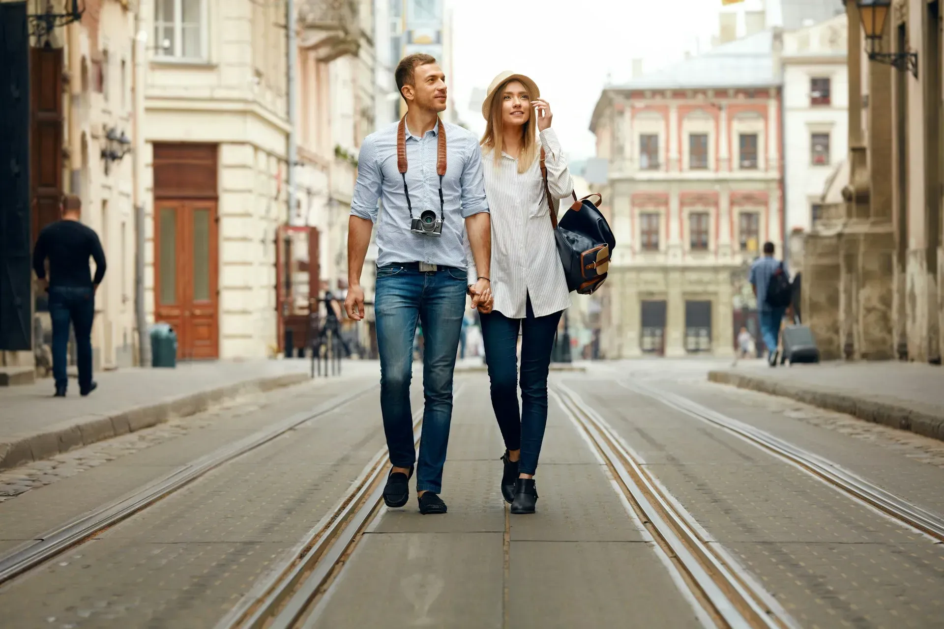 Couple walking hand-in-hand down a city street with vintage camera. Sunny day, European buildings.