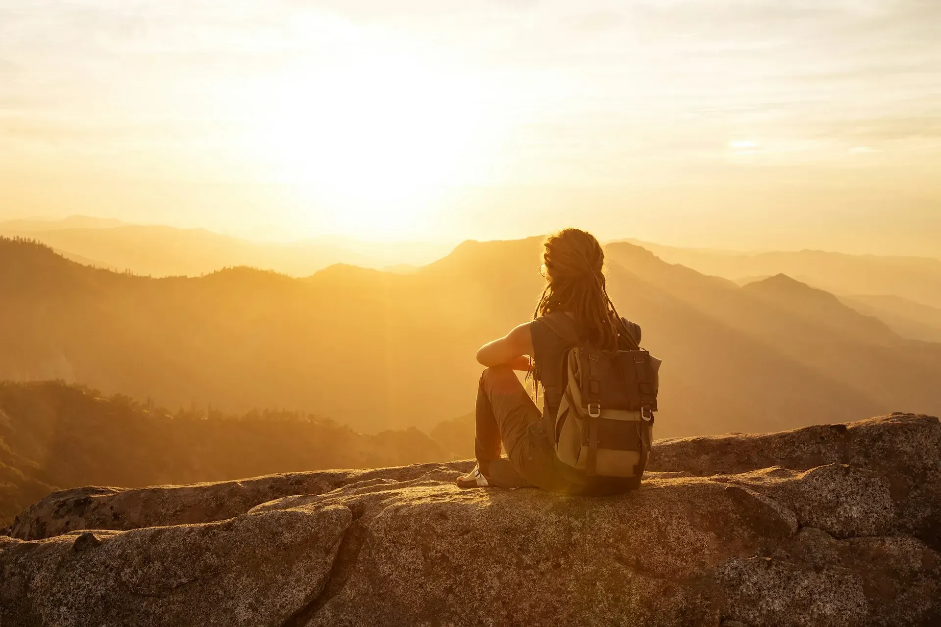 Person with backpack sits on a rock, watching the sunrise over mountains. Warm golden light.