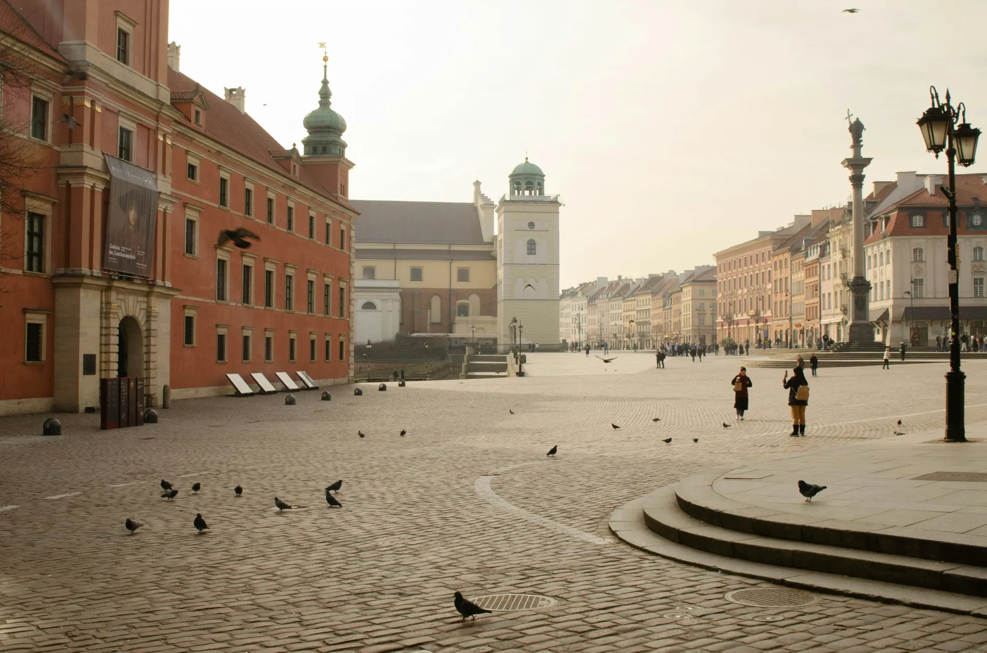 Cobblestone square in Warsaw, Poland, with red brick buildings, tower, and column. A few people and pigeons are present.