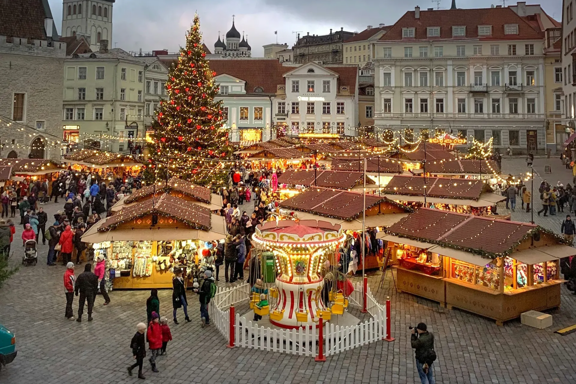 Christmas market in a European square with a large tree and carousel under a stormy sky.