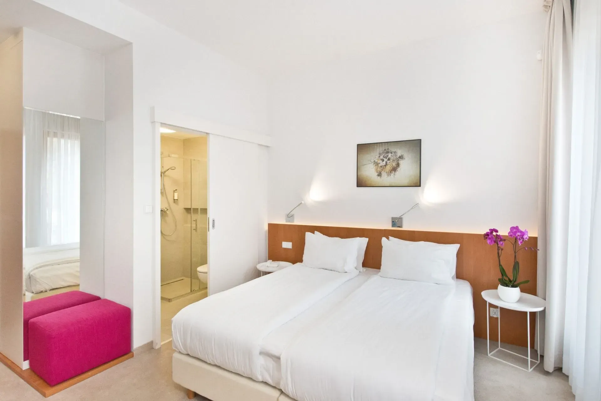 Jasna sypialnia hotelowa z białą pościelą, różową ławką, drewnianym zagłówkiem i kwiatami przy łóżku