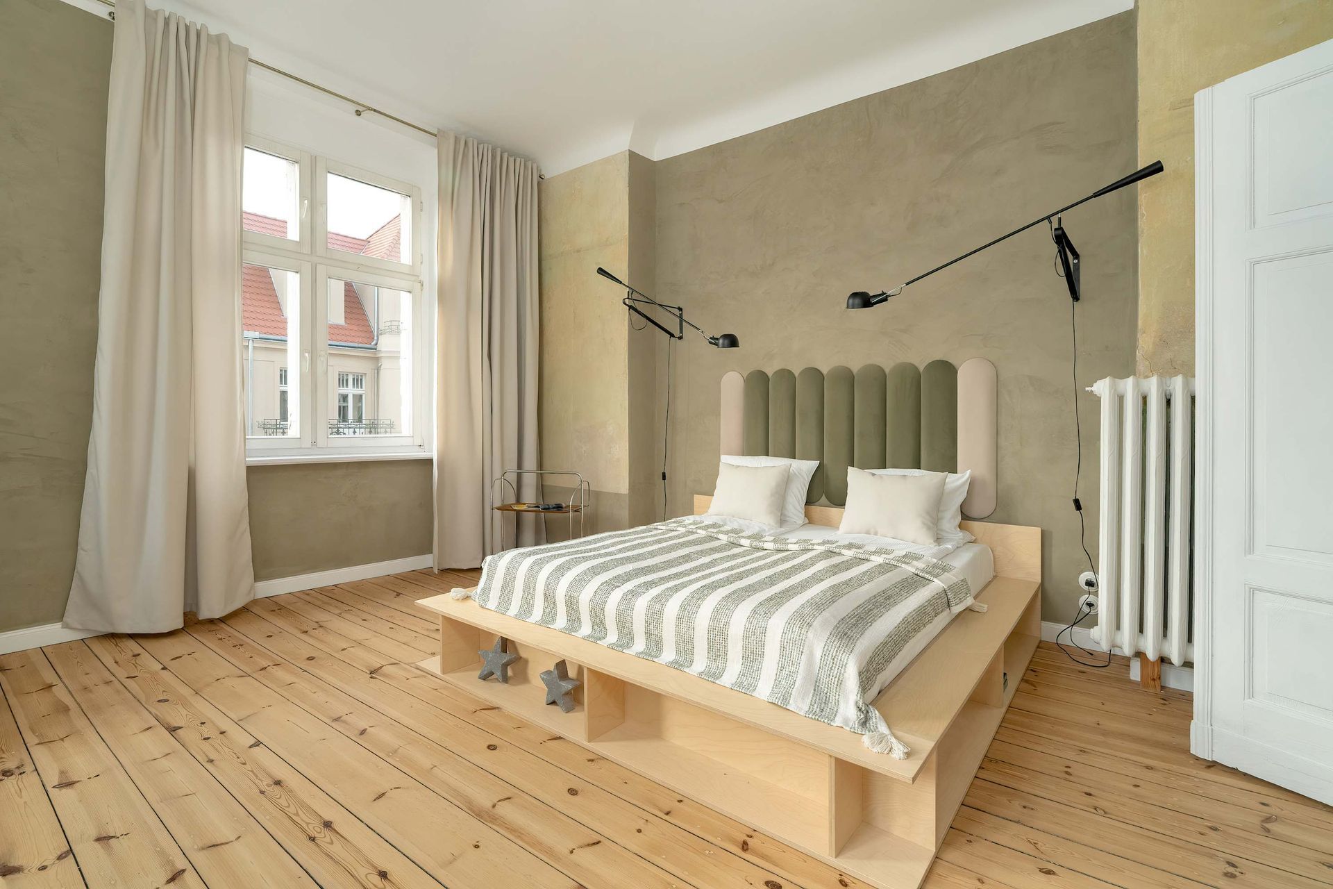 Sypialnia z jasną drewnianą podłogą, łóżkiem z zielonym zagłówkiem, oknem z zasłonami i kinkietami.