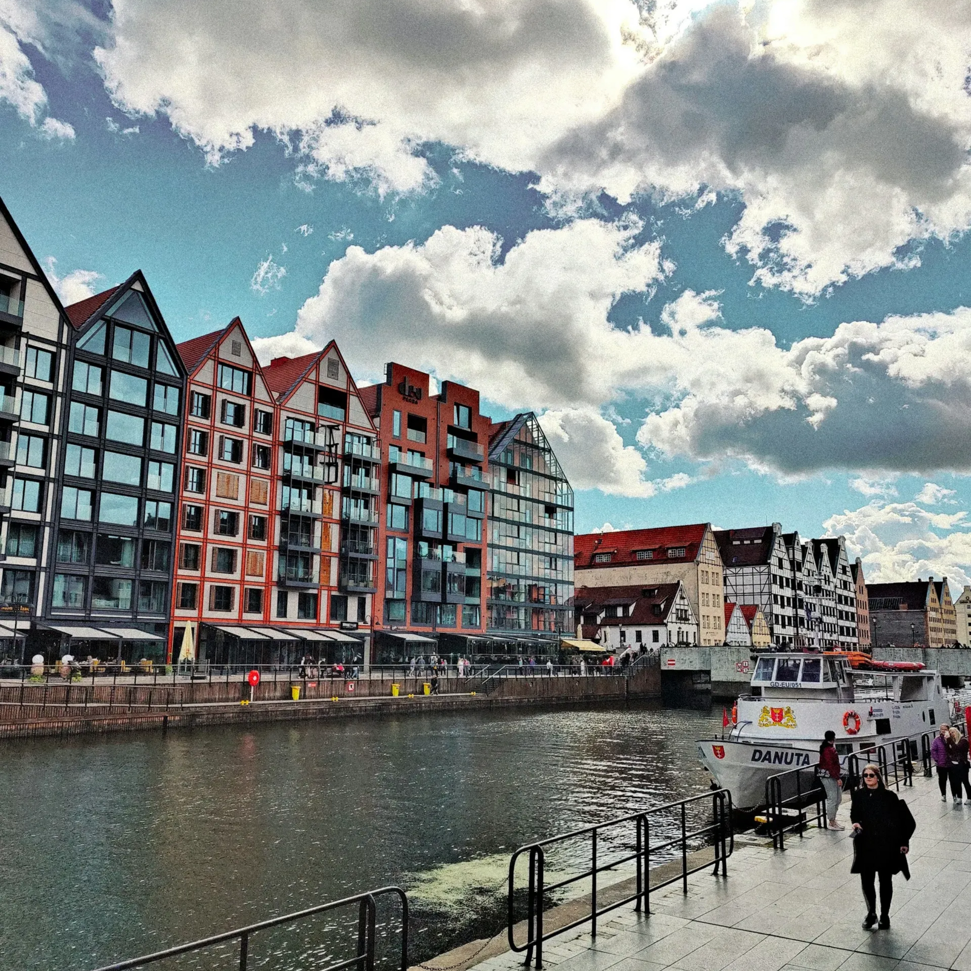 Budynki nadbrzeżne z czerwonymi i niebieskimi akcentami pod zachmurzonym niebem. Osoba idzie brukowaną ścieżką.