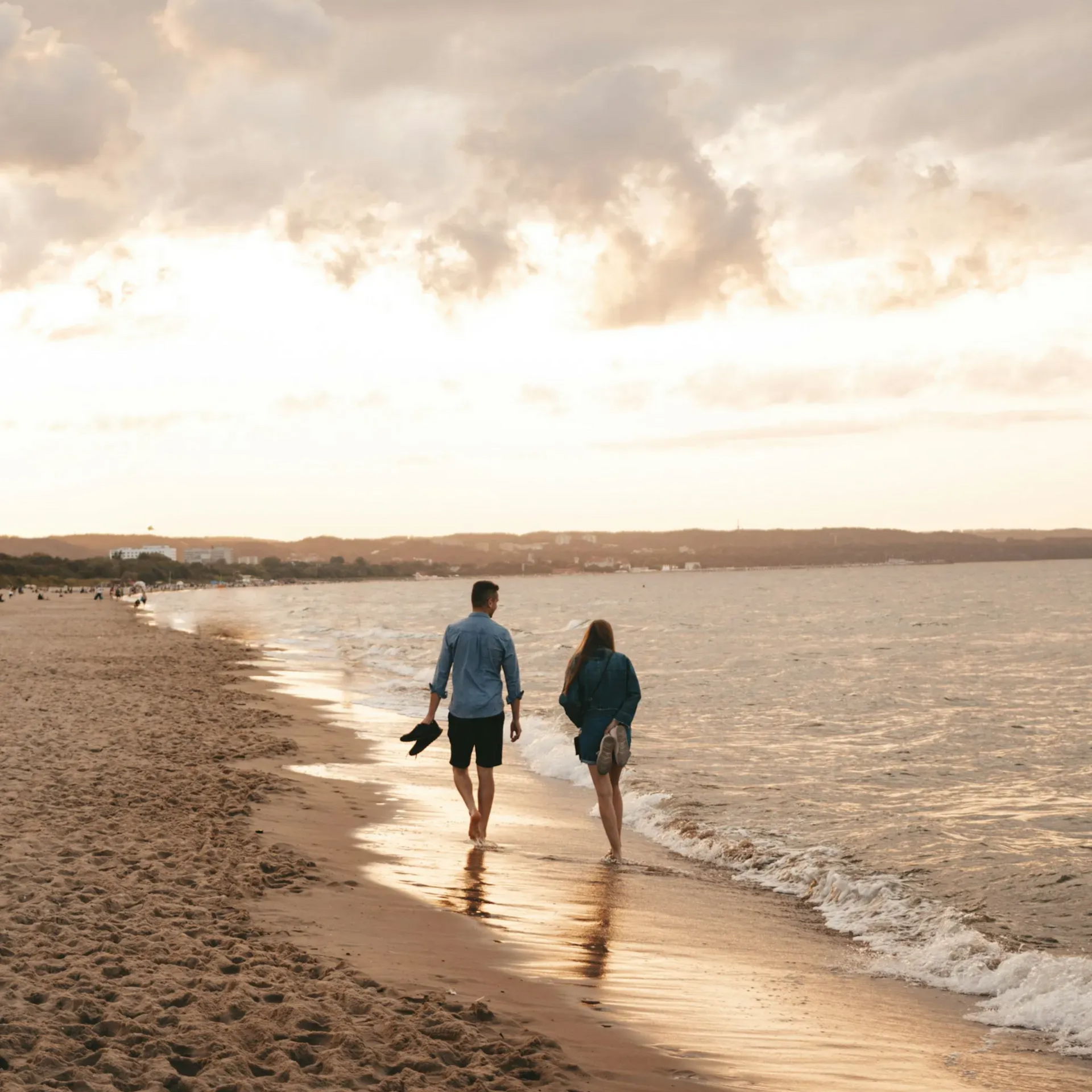 Para spaceruje po plaży o zachodzie słońca. Mężczyzna trzyma buty, kobieta ma na sobie kurtkę, a niebo jest zachmurzone.