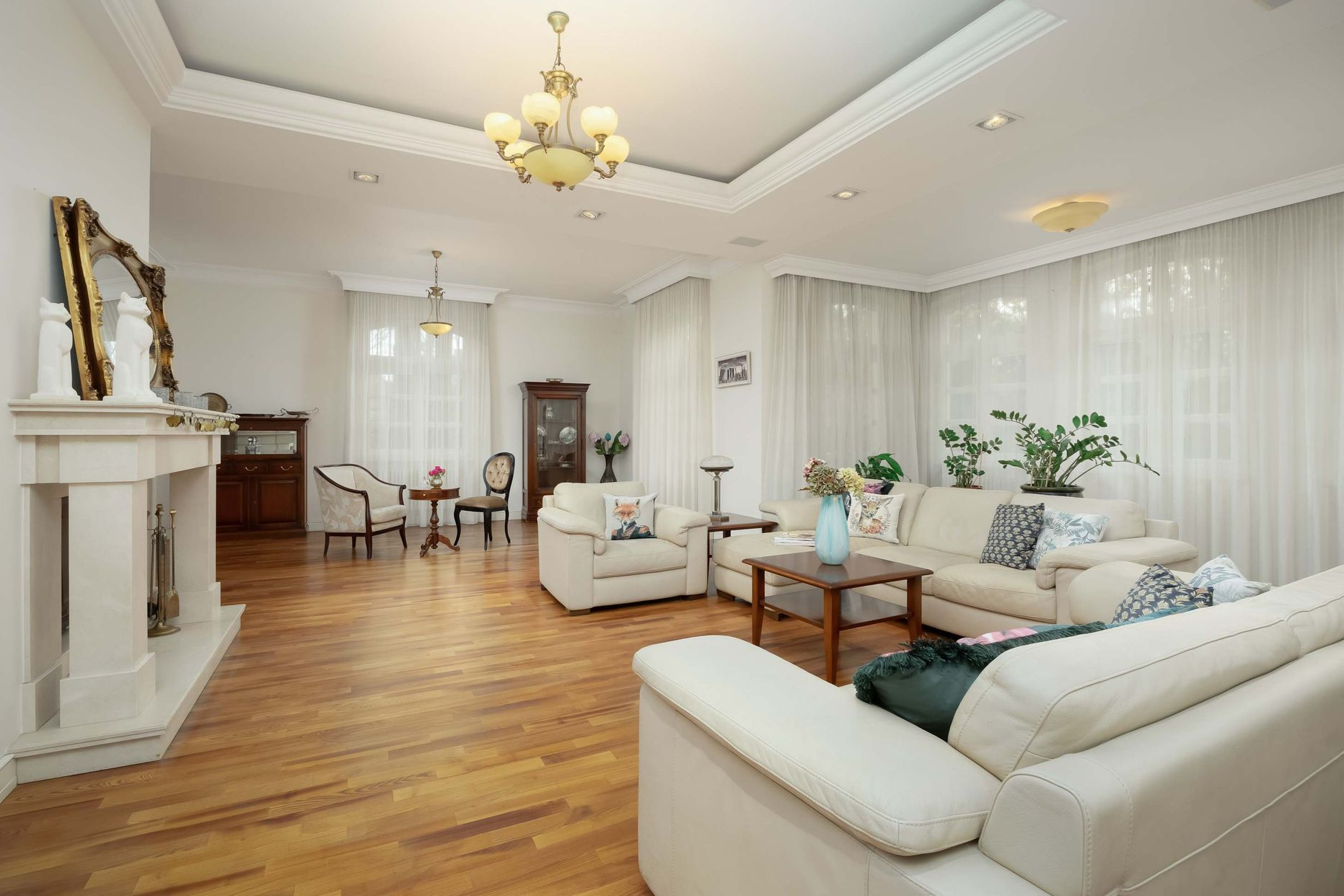 Przestronny salon z podłogą z twardego drewna, białymi meblami i dużymi oknami z delikatnymi zasłonami.