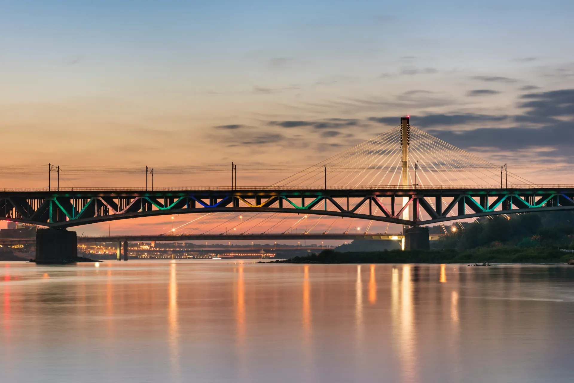 Bridge over water at sunset; illuminated in colors of teal, orange, and yellow; city in the background.