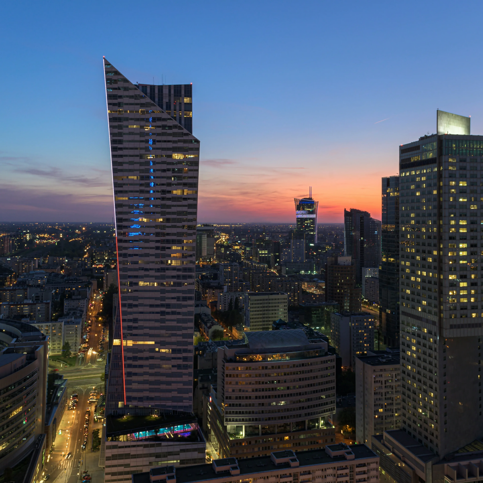 Panorama miasta o zmierzchu, z oświetlonymi wieżowcami na tle kolorowego nieba.