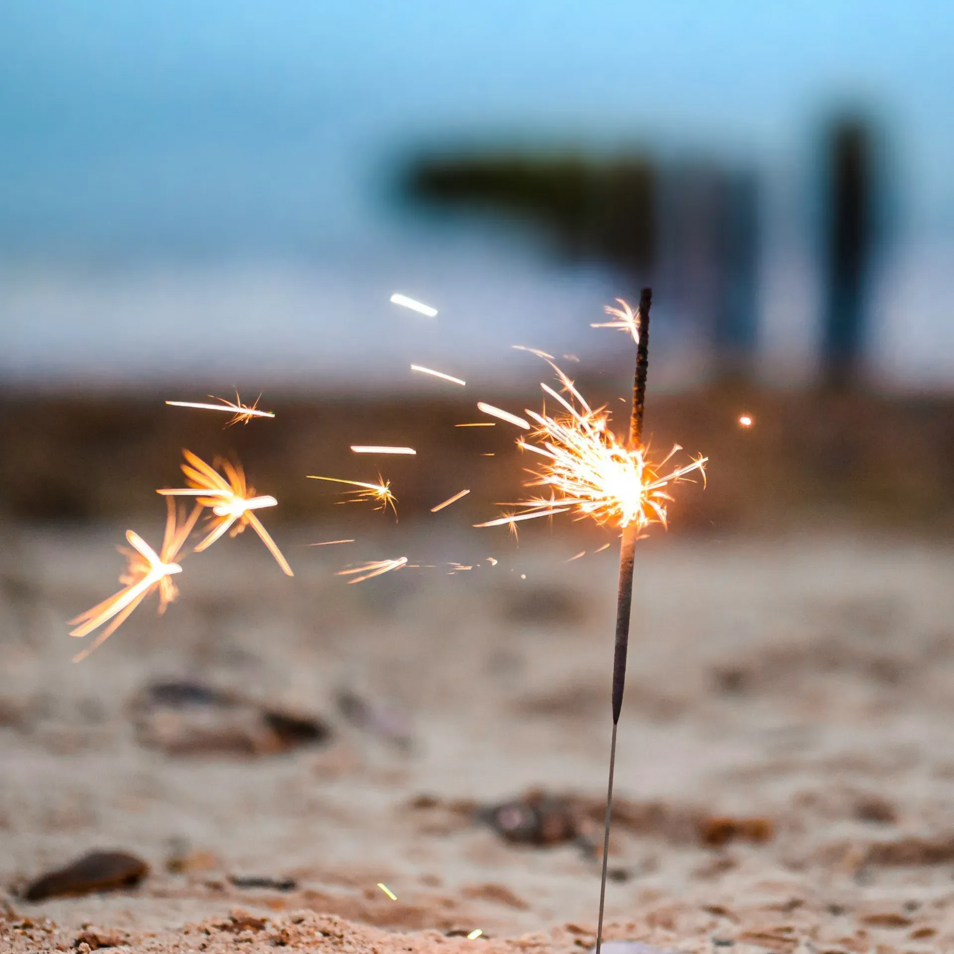 Zimne ognie emitujące iskry na piaszczystej plaży, rozmazane postacie w tle, błękitne niebo.