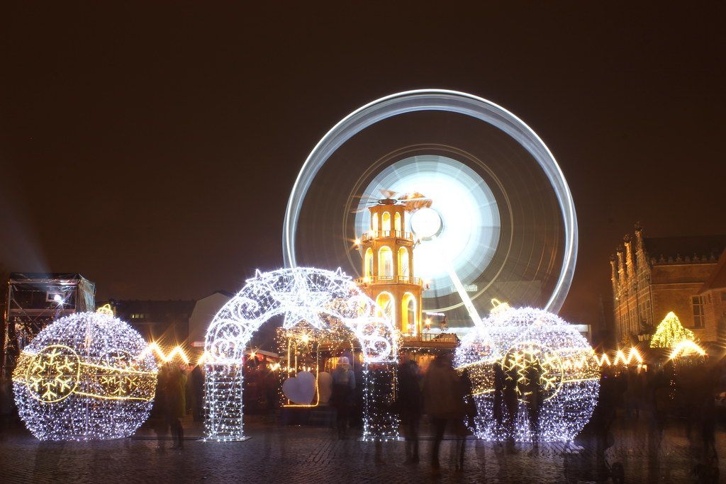 Night scene of a Christmas market with illuminated decorations, Ferris wheel, and crowds of people.
