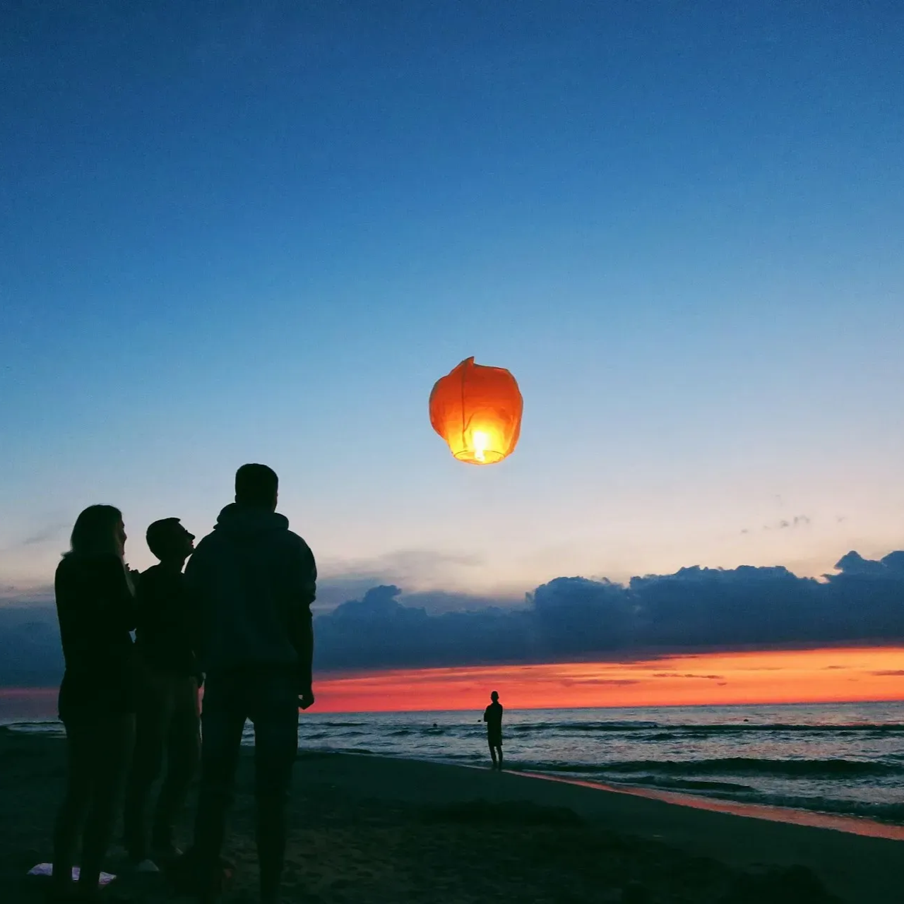Ludzie na plaży obserwują, jak zapalony papierowy lampion wznosi się na niebo nad oceanem o zmierzchu.