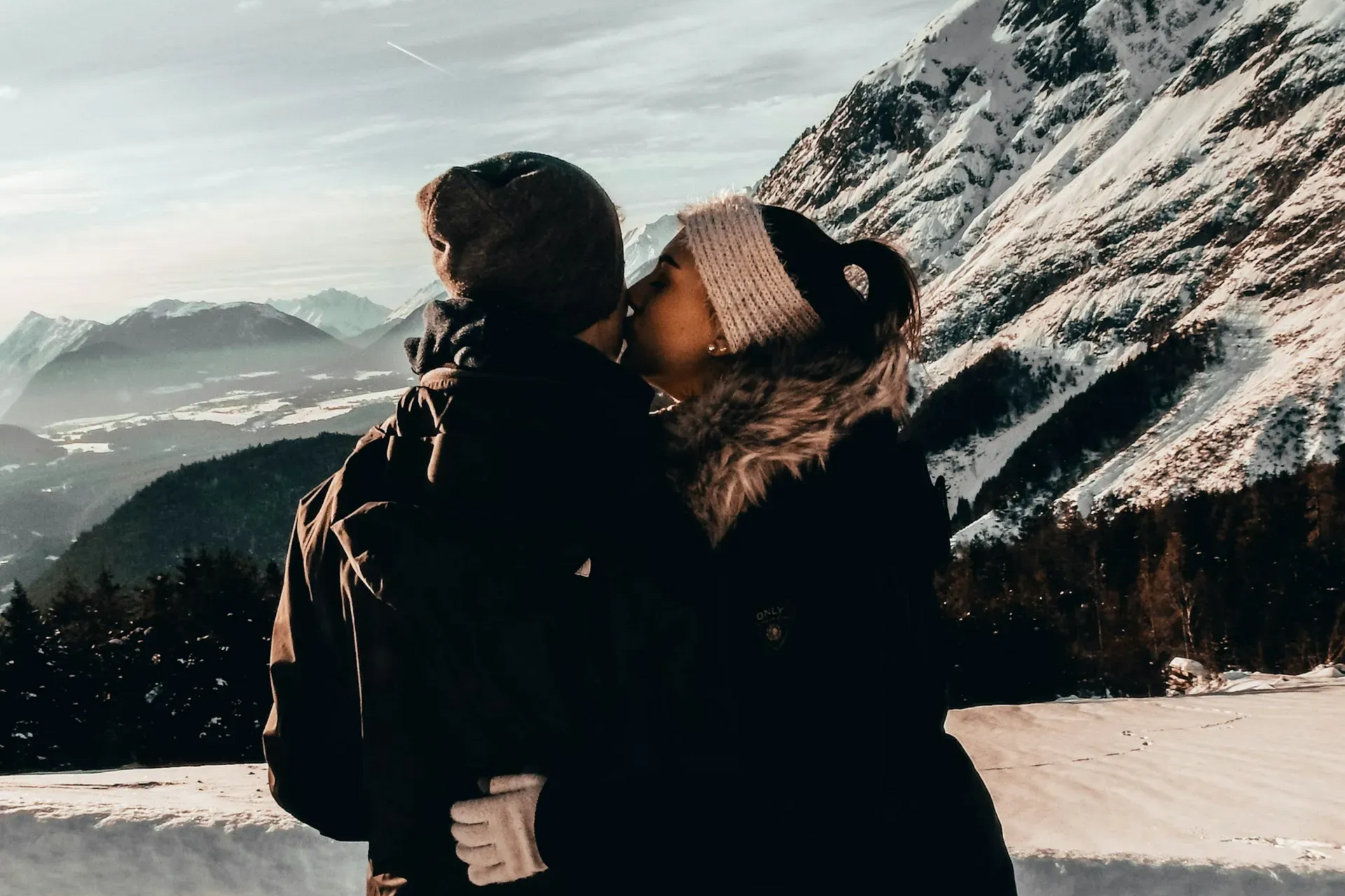 Couple kissing in snowy mountains; both wearing winter coats and hats.