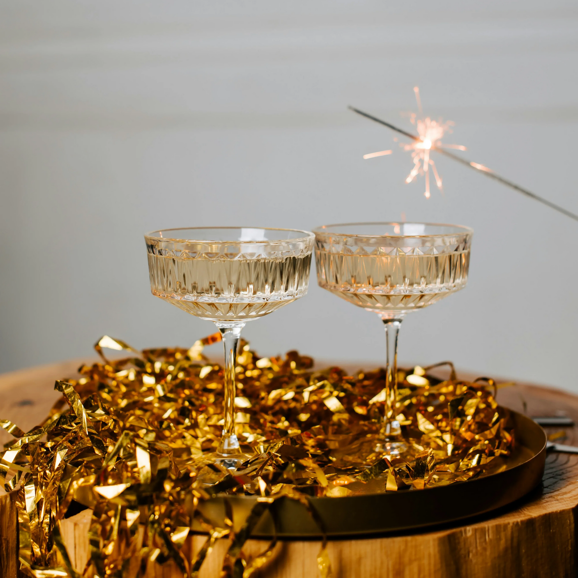 Two champagne glasses on a tray with gold tinsel, a lit sparkler in background.