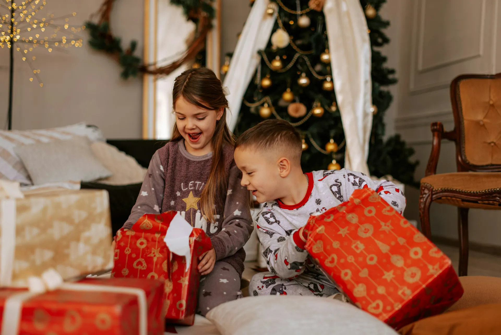 Two children opening Christmas presents in front of a tree. The girl is surprised, boy is excited. Room is decorated.