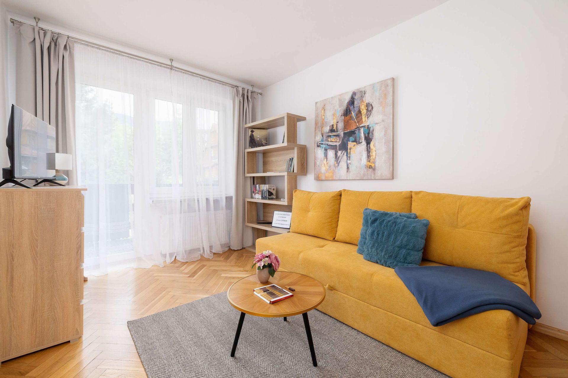 Jasny salon z żółtą sofą, stolikiem kawowym, dywanem, regałem na książki i dużym oknem z cienkimi zasłonami