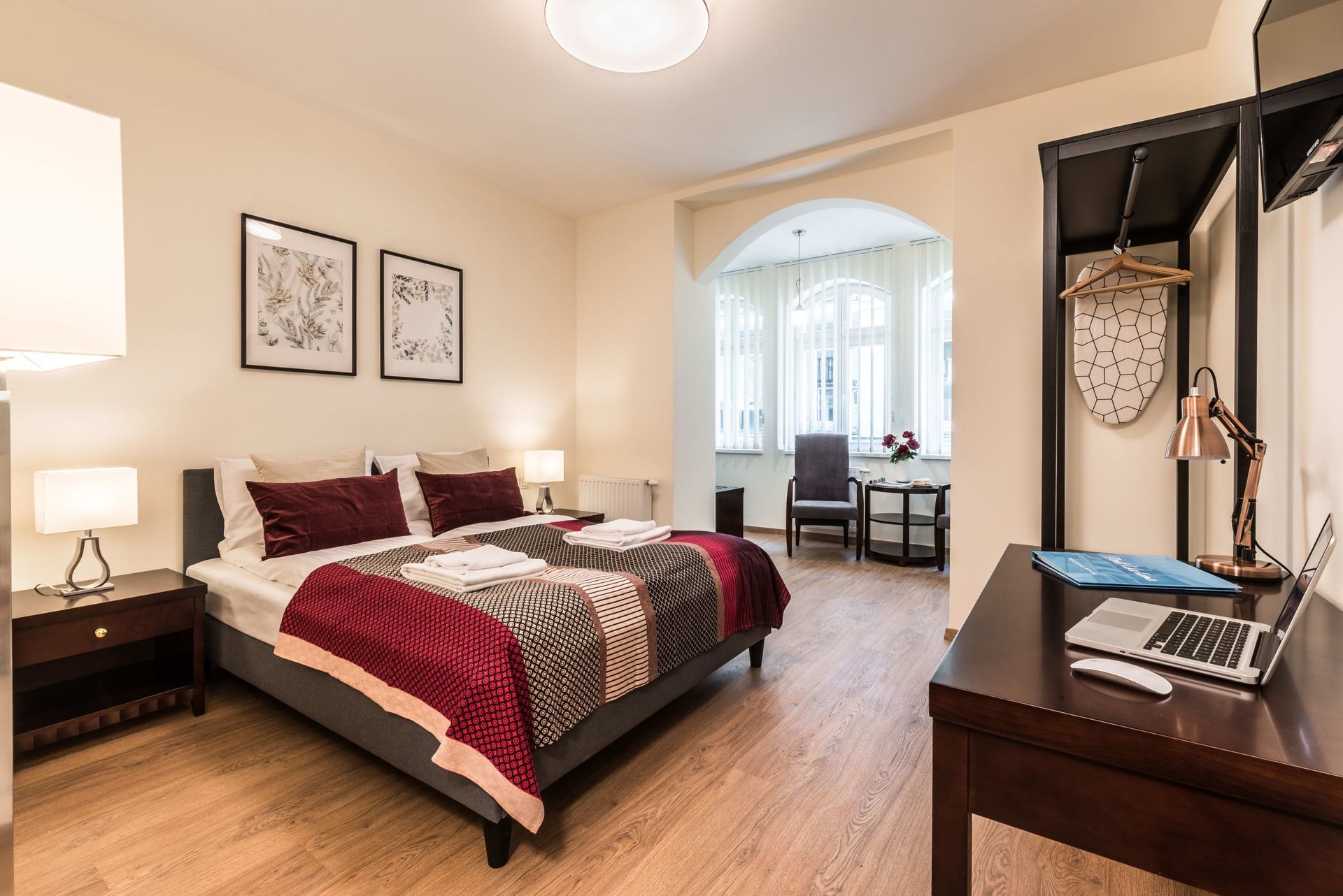 Sypialnia z łóżkiem podwójnym, biurkiem z laptopem i małym kącikiem wypoczynkowym przy oknie.