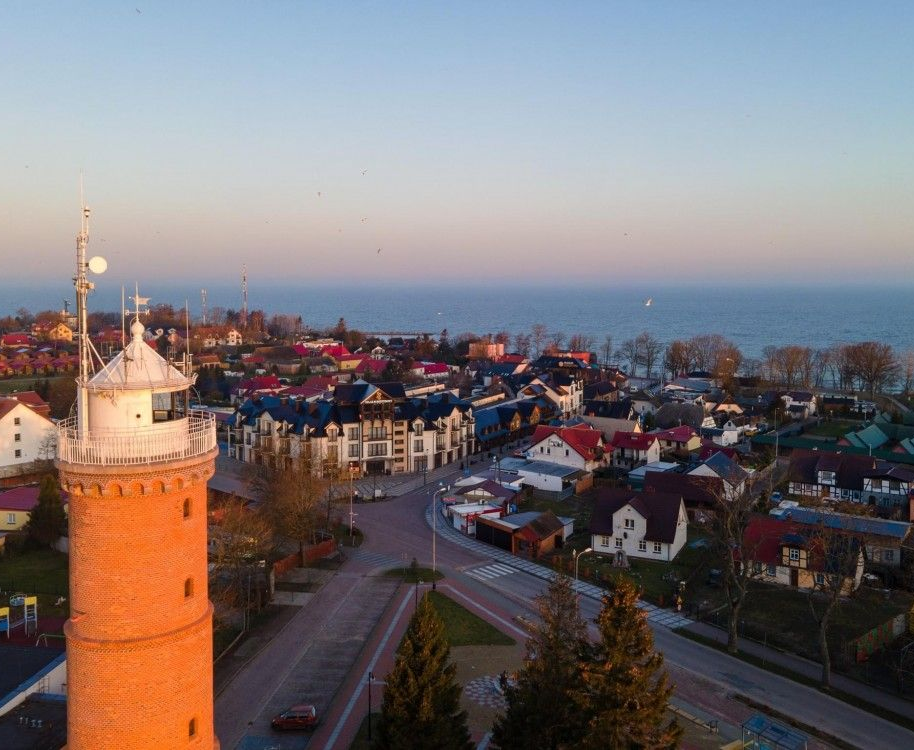 Widok z lotu ptaka na nadmorskie miasteczko z czerwoną ceglaną latarnią morską, budynkami, drogą i morzem pod czystym niebem.