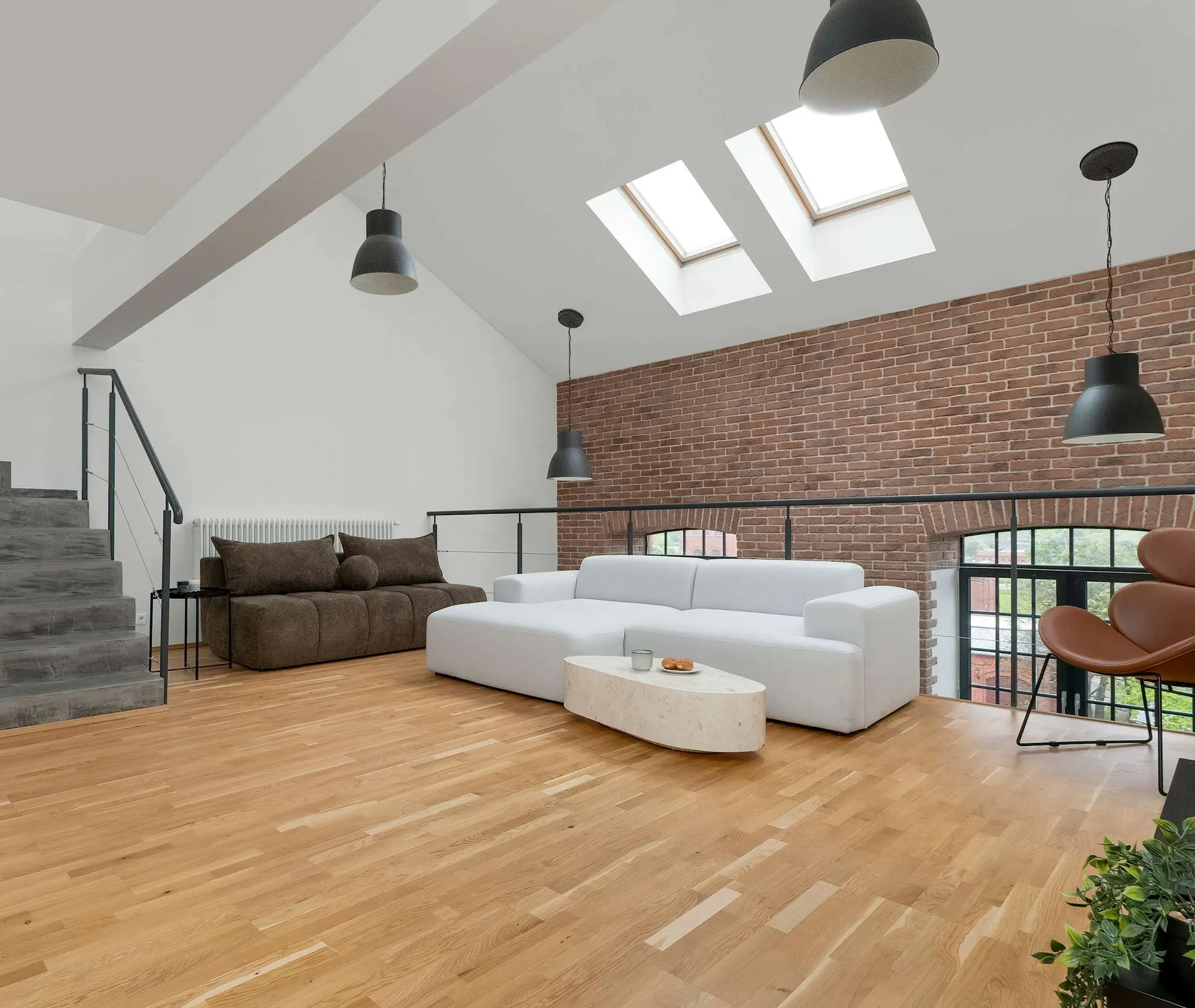 Salon w stylu loft z białą, narożną sofą, odsłoniętą ceglaną ścianą, świetlikami i drewnianą podłogą.