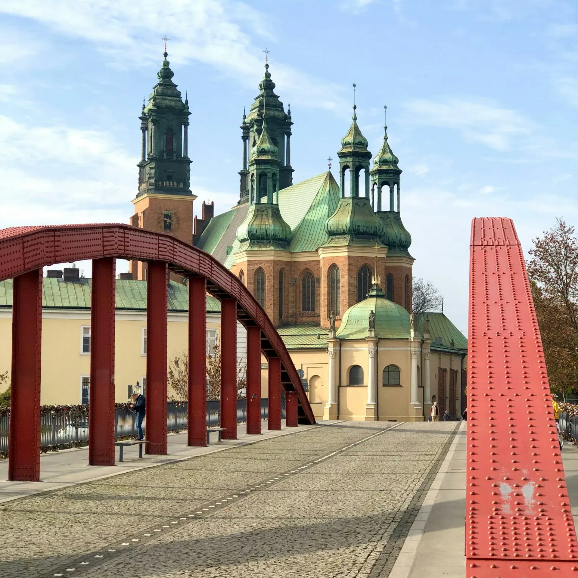 Czerwony łukowy most z brukowaną nawierzchnią prowadzi do katedry z zielonymi kopułami i wieżami w Poznaniu, w Polsce.