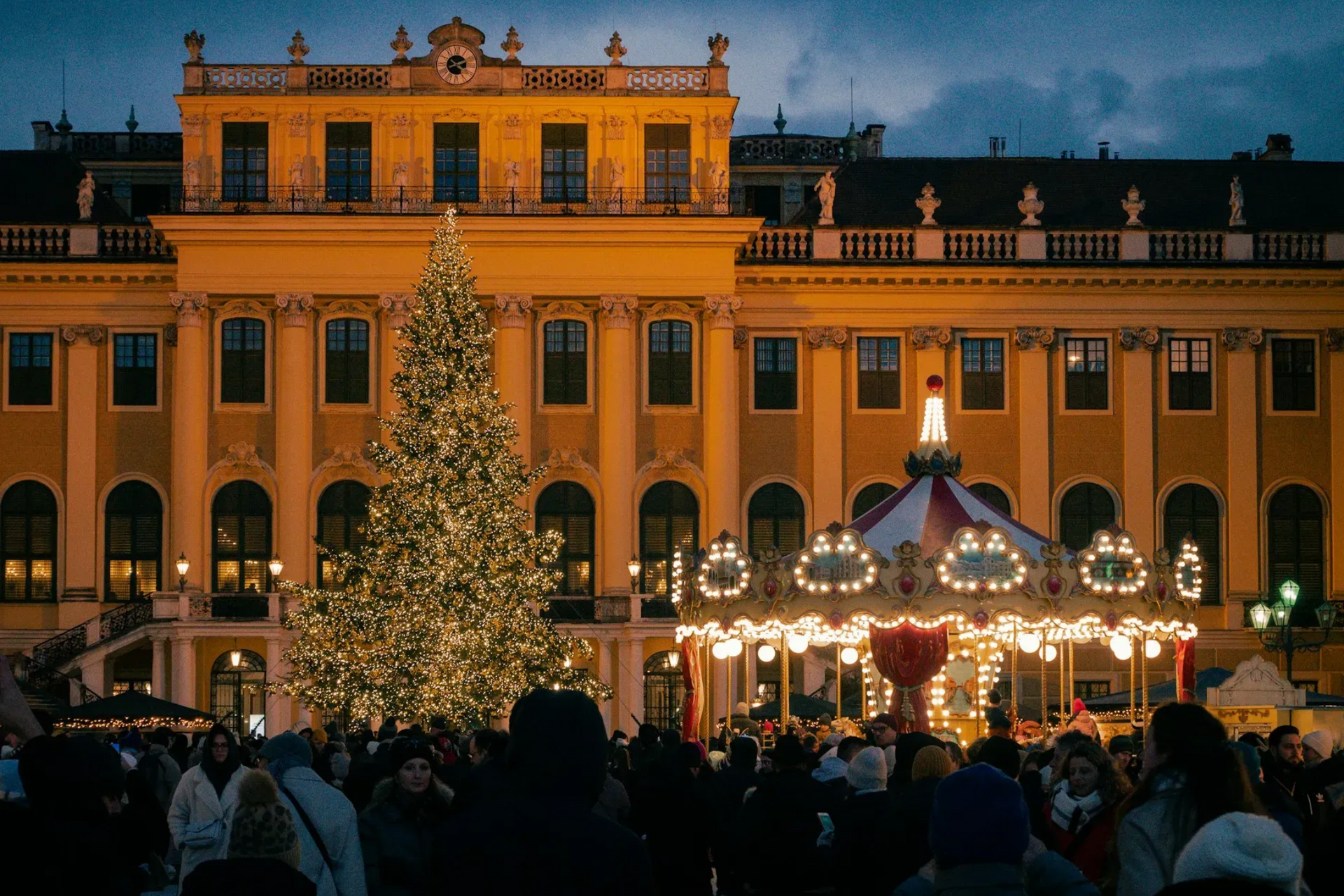 Christmas market with a large decorated tree and carousel in front of a golden-lit palace. People are visible.