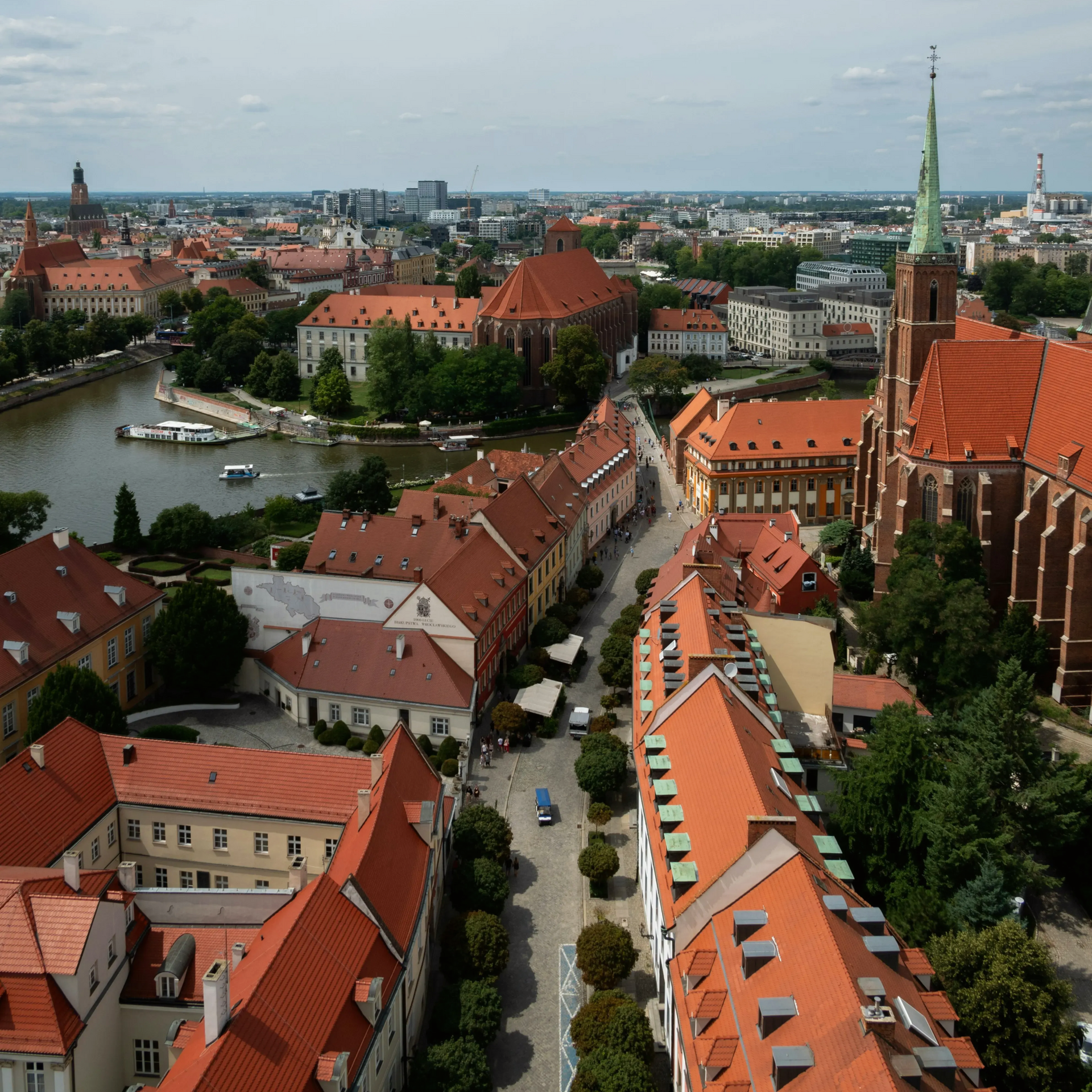 Aerial view of a Wroclaw city with red-tiled roofs, a river, and a tall church spire under a cloudy sky.