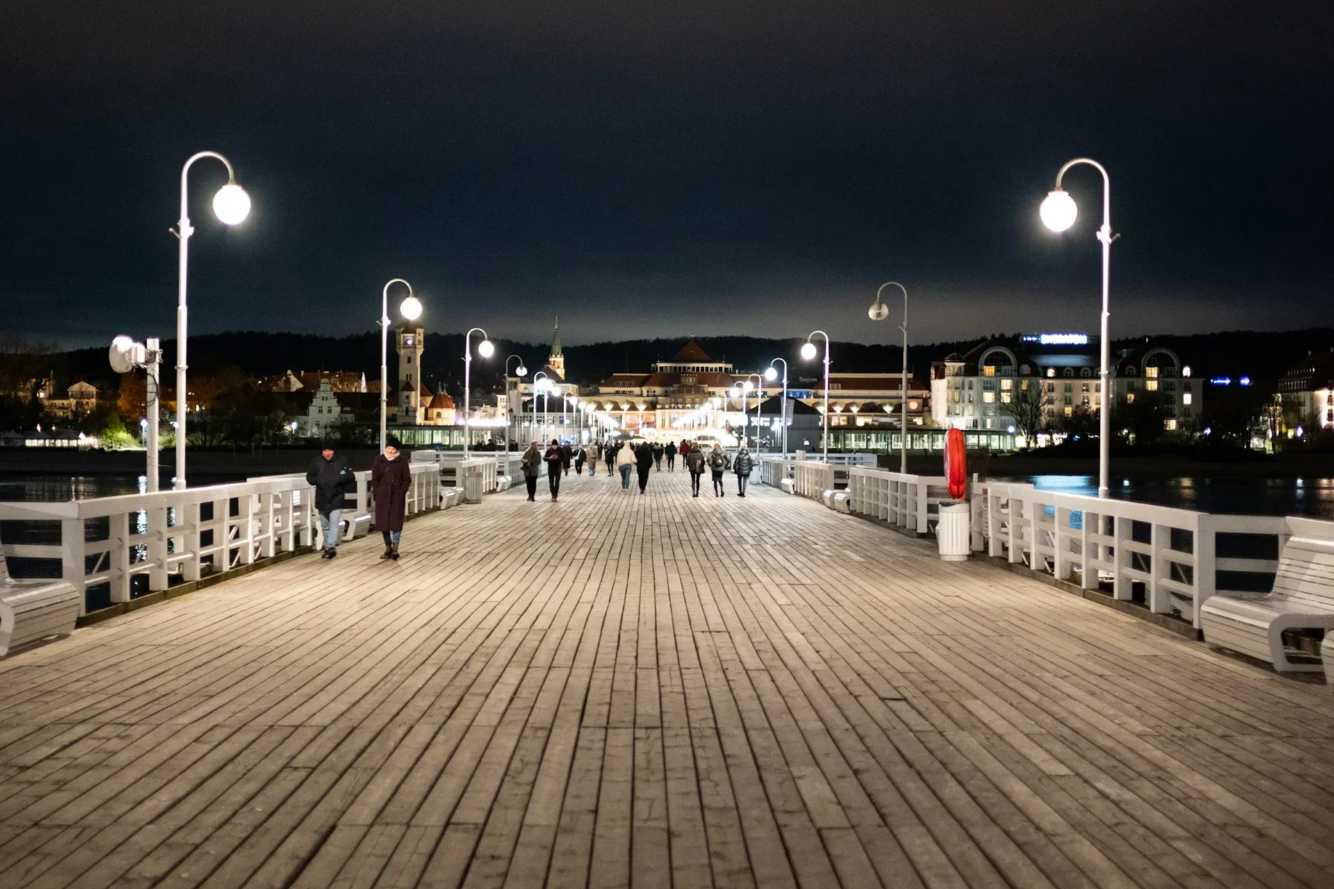 Wooden pier at night with people walking towards city lights. Lit by white street lamps.