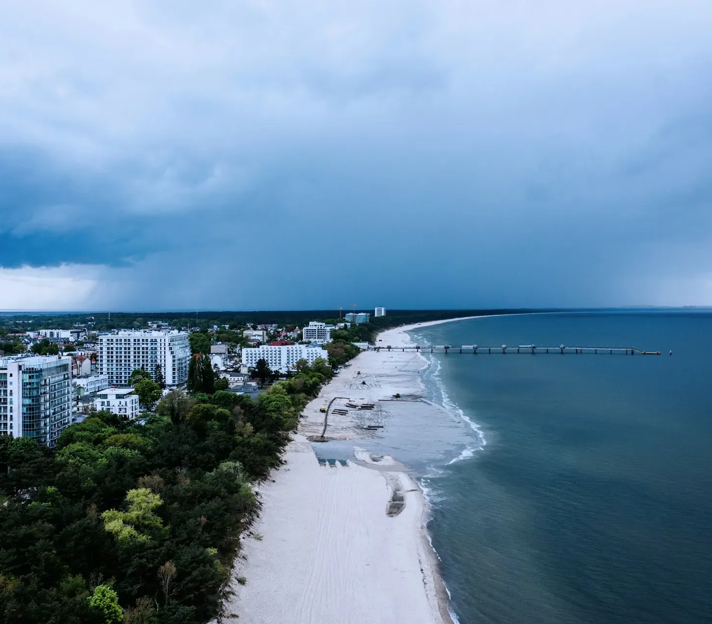 Widok na plażę pod zachmurzonym niebem, z budynkami, drzewami i molo wzdłuż linii brzegowej.
