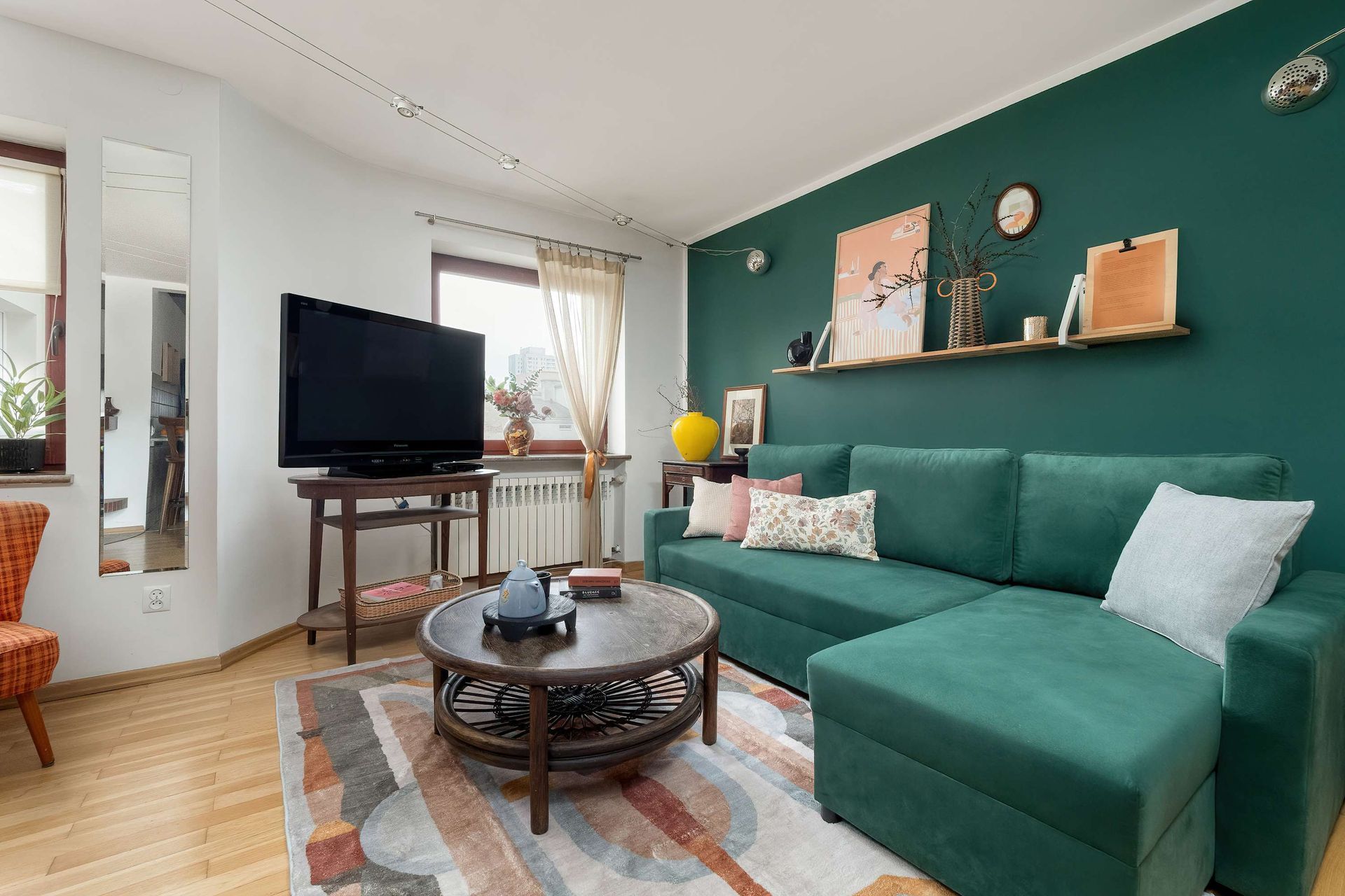 Salon z zieloną sofą, turkusową ścianą, telewizorem i wzorzystym dywanem.