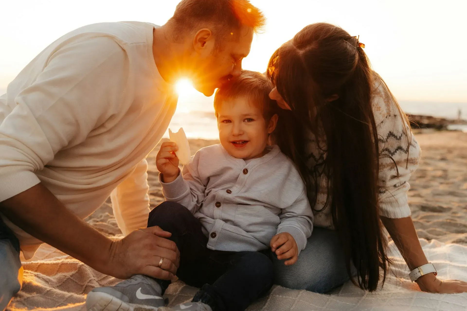 Family of three at beach with sunset: parents kissing child on head, all smiling.