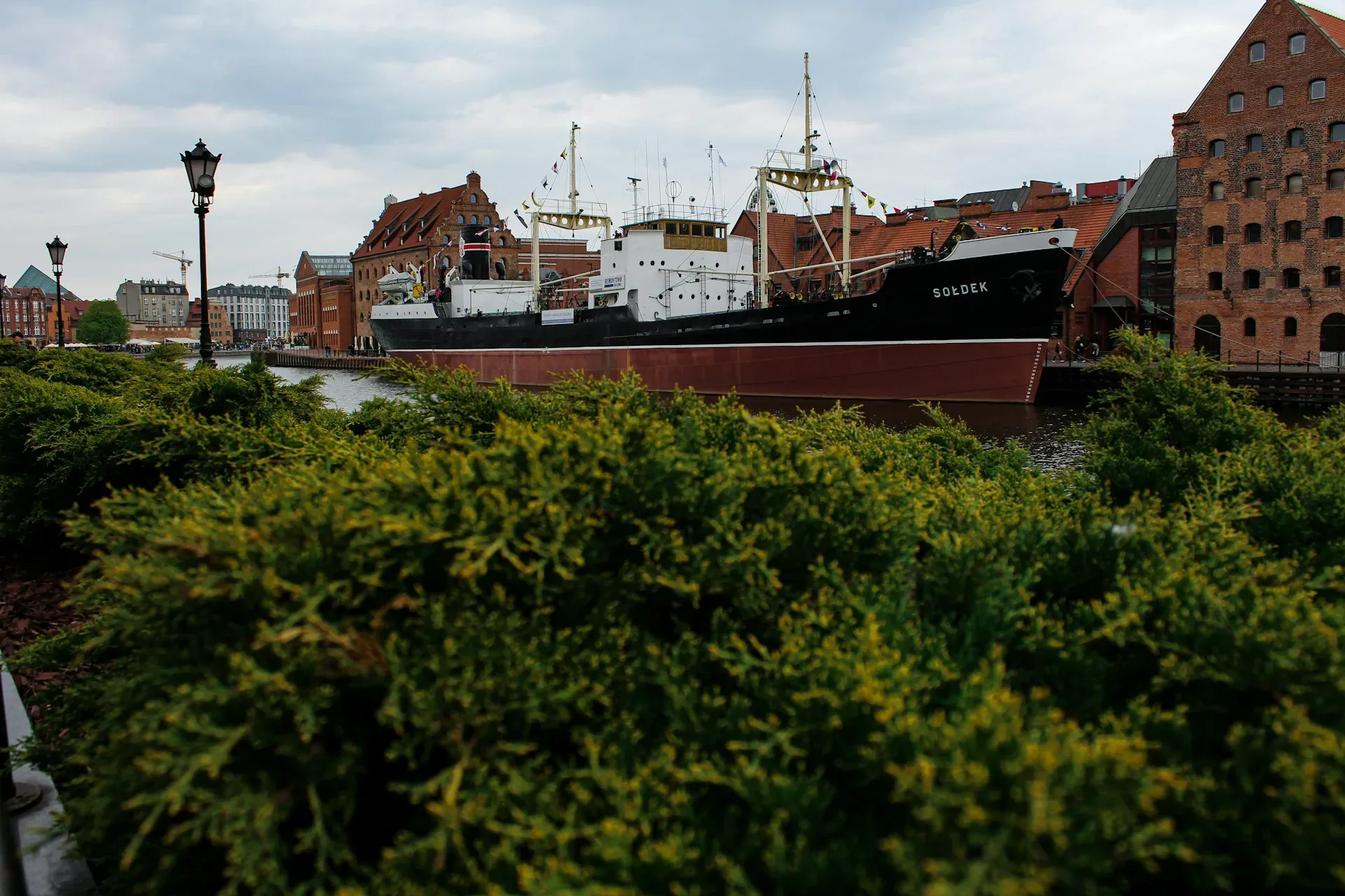 Green bushes in the foreground, a large ship docked in a river, brick buildings in the background.