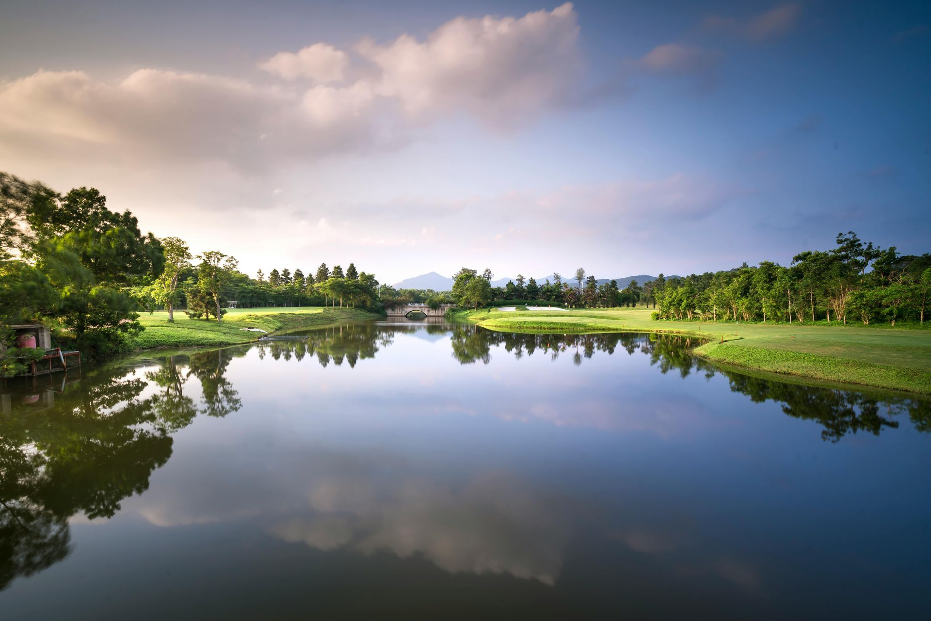 Spokojne jezioro odbija zachmurzone niebo i zielone pole golfowe otoczone drzewami.