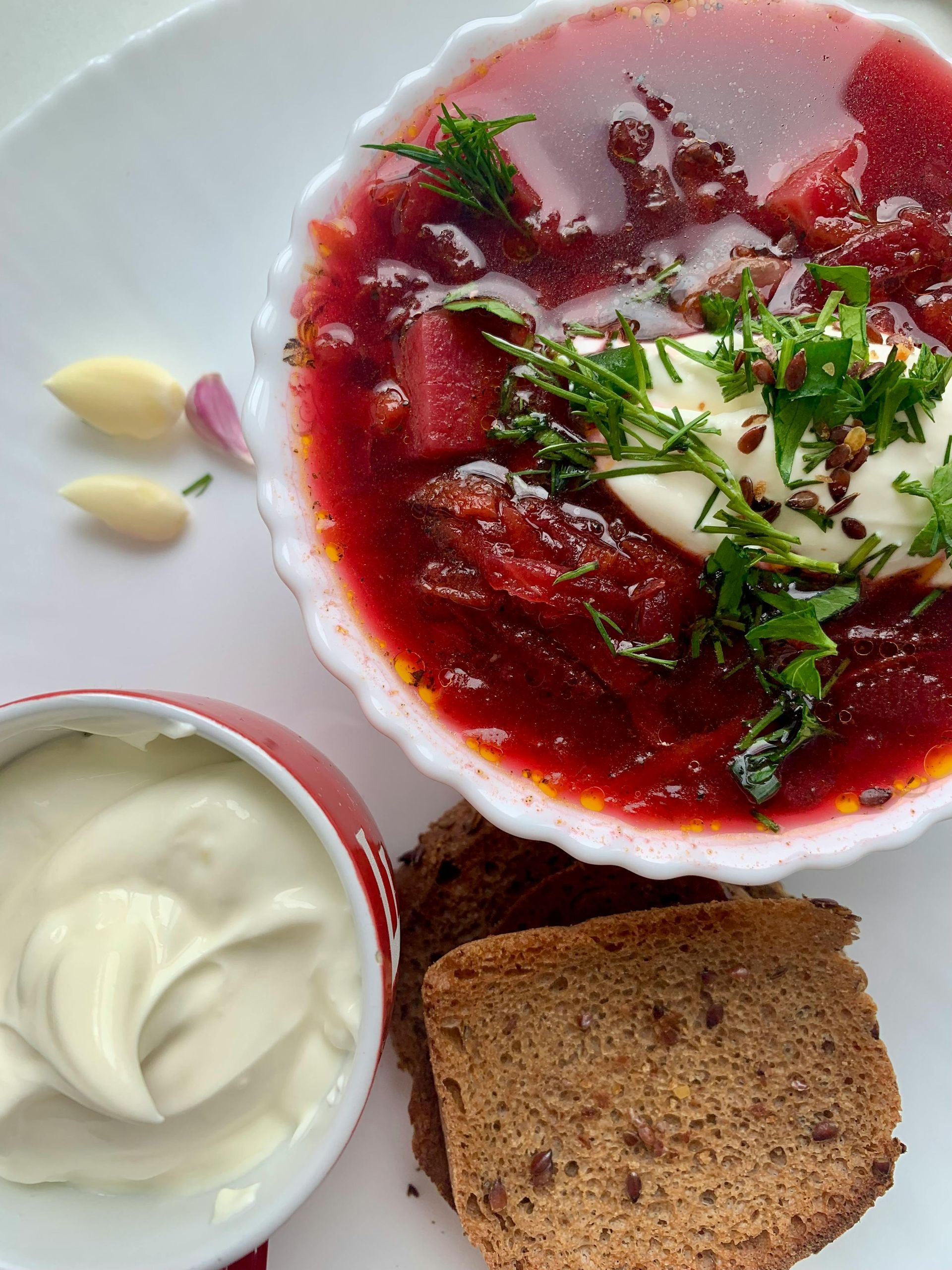 Bowl of red borscht, topped with herbs and sour cream, served with bread and cream on a white plate.