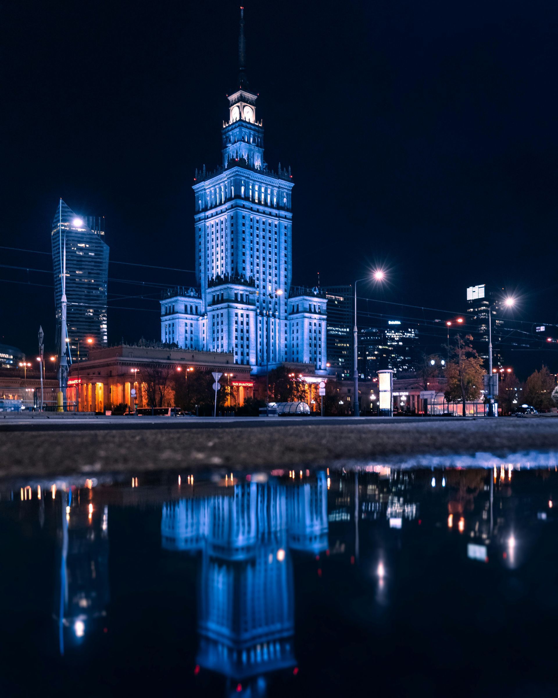 Nocny widok Warszawy, Polska: Pałac Kultury podświetlony na niebiesko, odbijający się w wodzie.