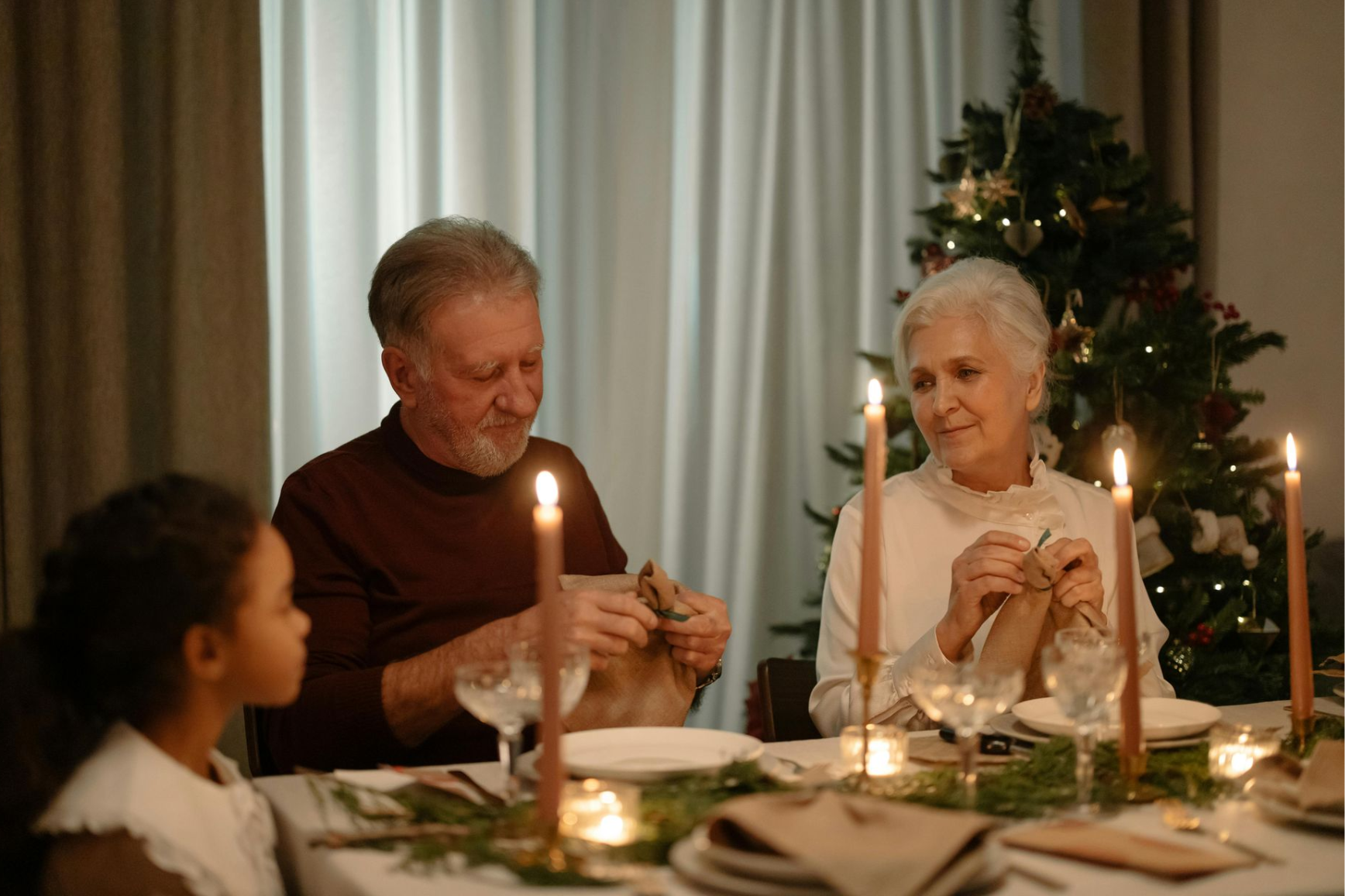Family at a Christmas table opening gifts, lit by candles and a decorated tree.